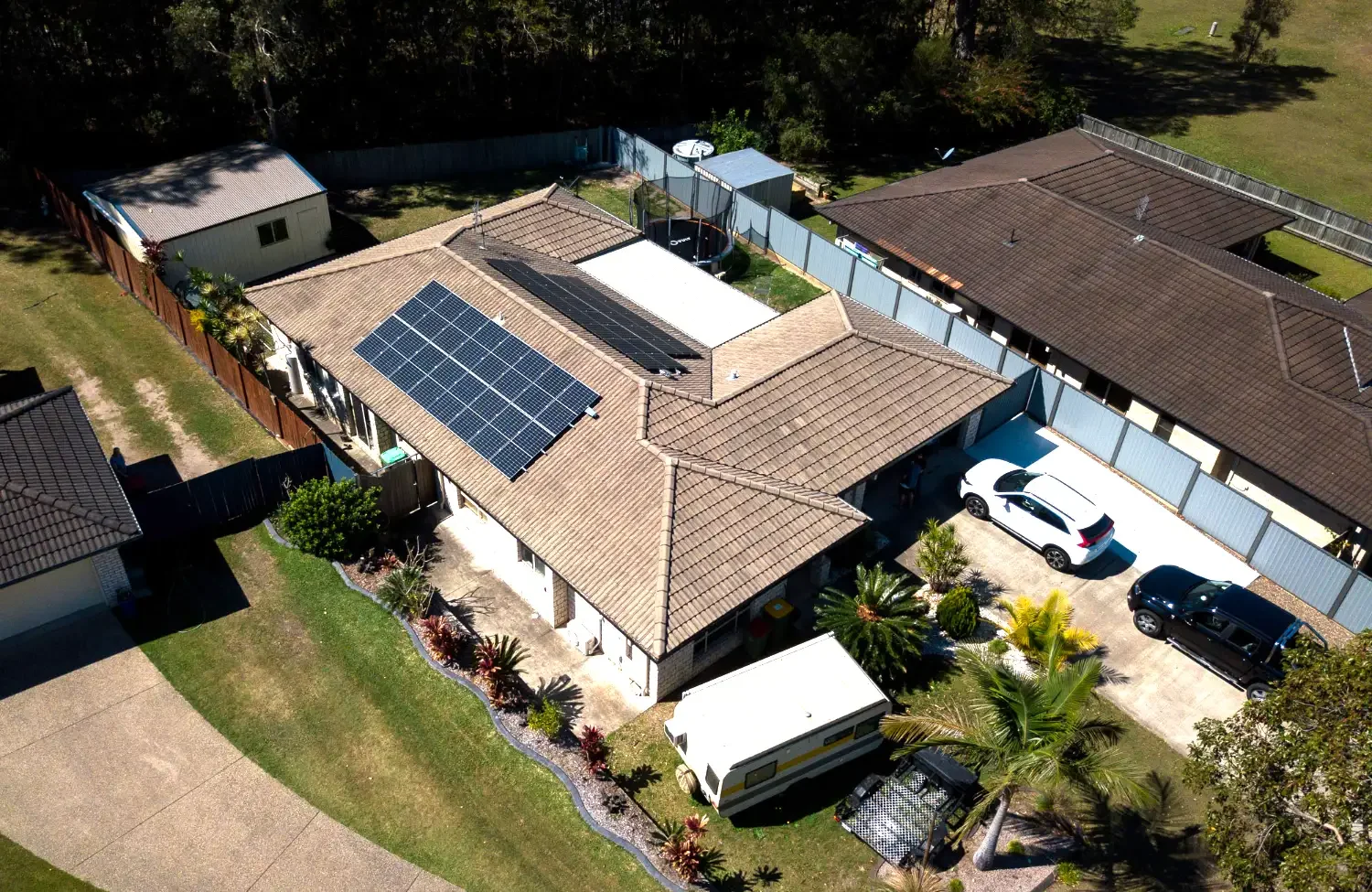 Aerial view of a house with solar panels on the roof, a backyard with a trampoline, a small shed, a caravan, and a fenced yard surrounded by neighboring houses and trees.