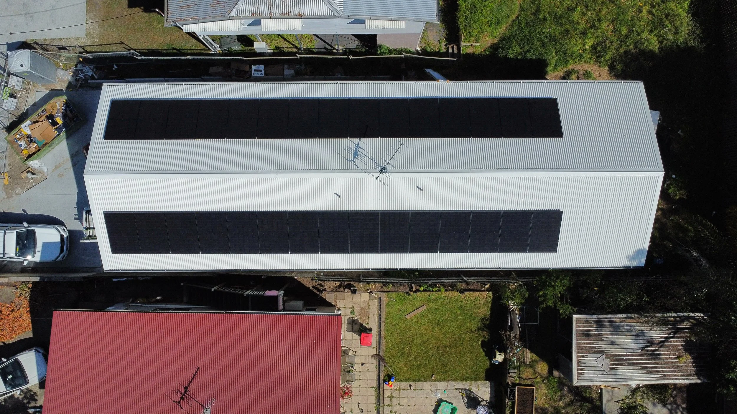 Aerial view of a large white metal-roof building with solar panels on top, adjacent to a smaller red-roof building and a backyard with toys and furniture.