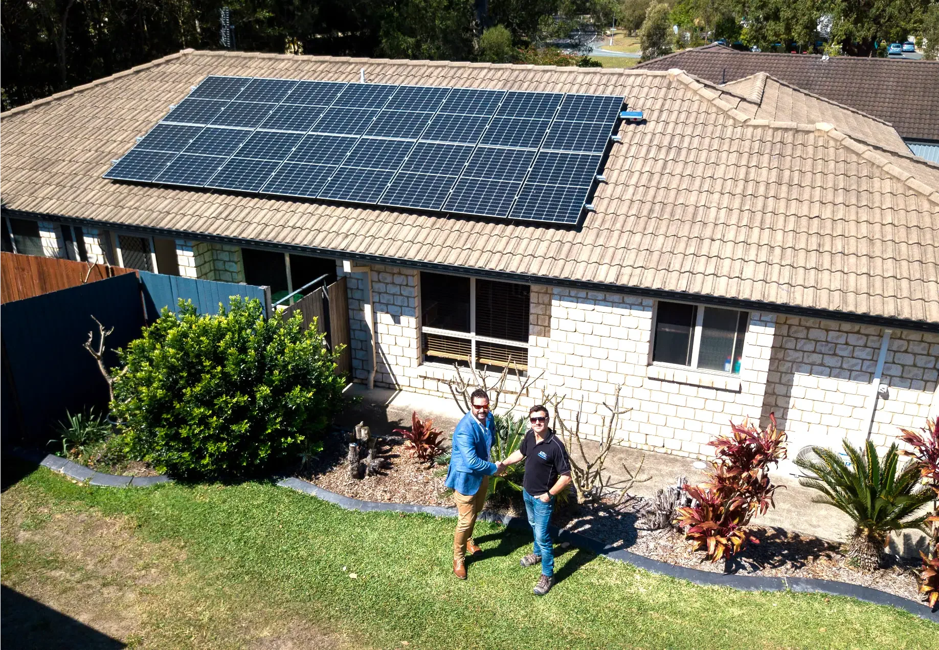 Two men shake hands in front of a house with solar panels on the roof, surrounded by a garden with trees and plants.