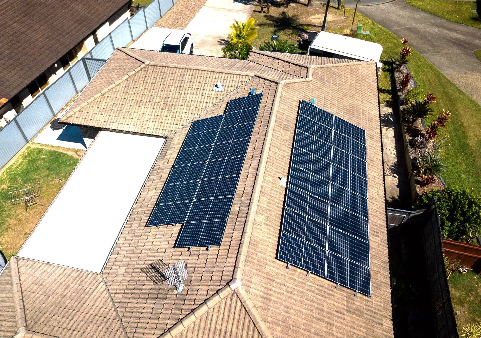 Aerial view of a house with two large solar panel arrays on its brown tiled roof, surrounded by a fenced yard, other houses, a driveway with parked cars, and a garden with various plants.