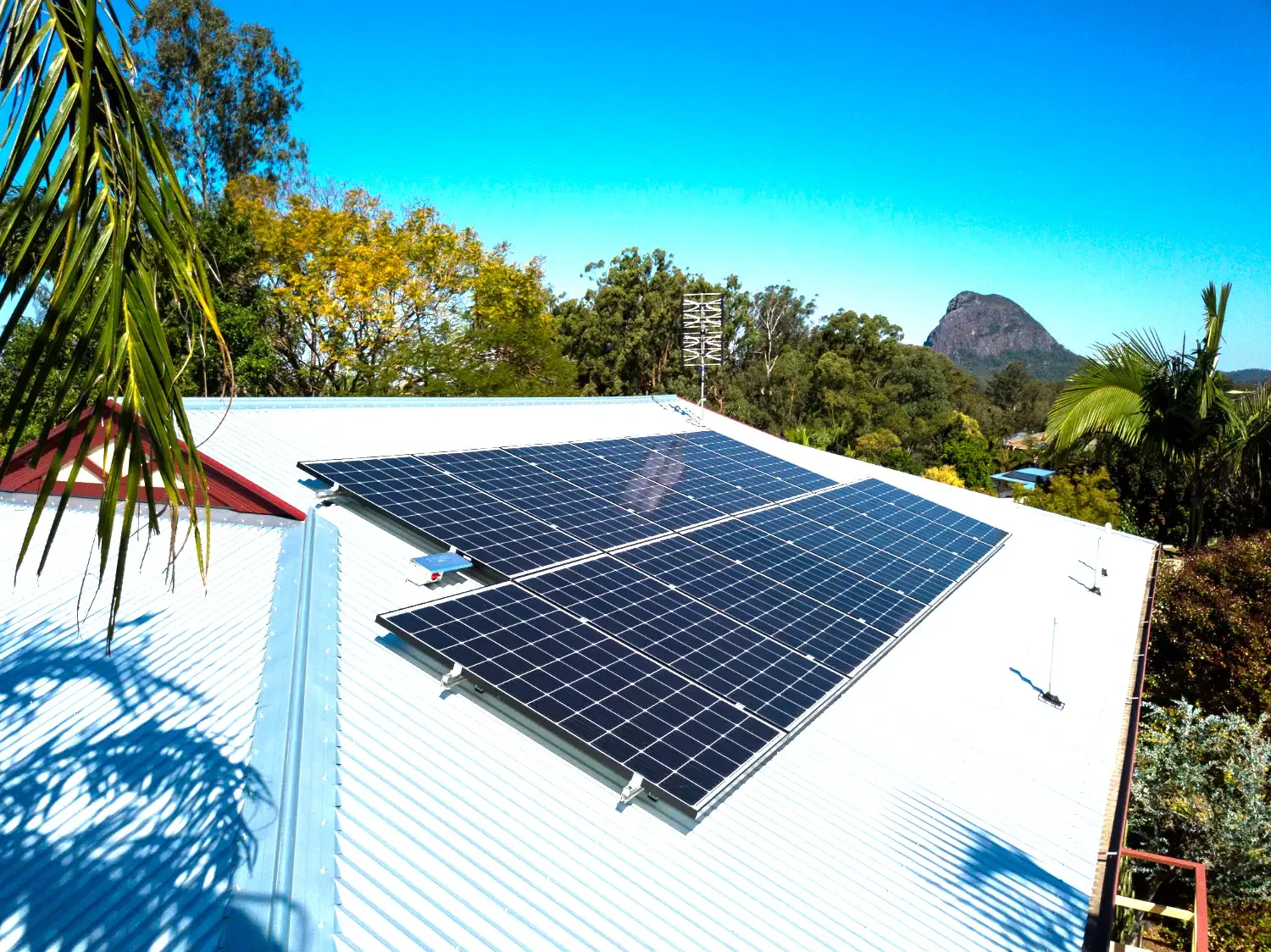Solar panels installed on a white metal rooftop with trees and mountain in the background under a clear blue sky.