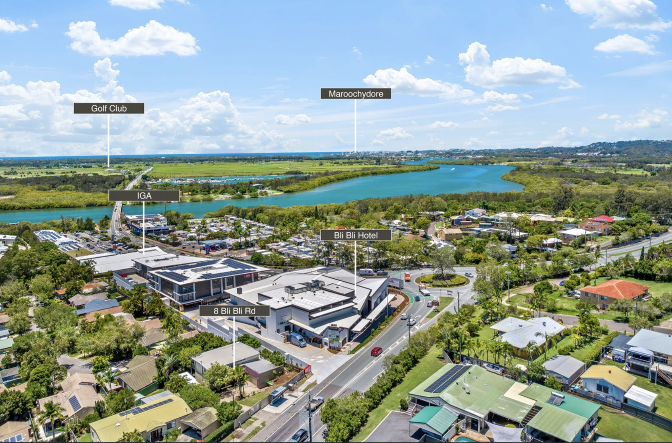 Aerial view of a city near a river with labeled landmarks, including a golf club, a hotel called Bli Bli Hotel, an IGA supermarket, and a road named 8 Bli Bli Rd, surrounded by houses, trees, and green fields under a partly cloudy sky.