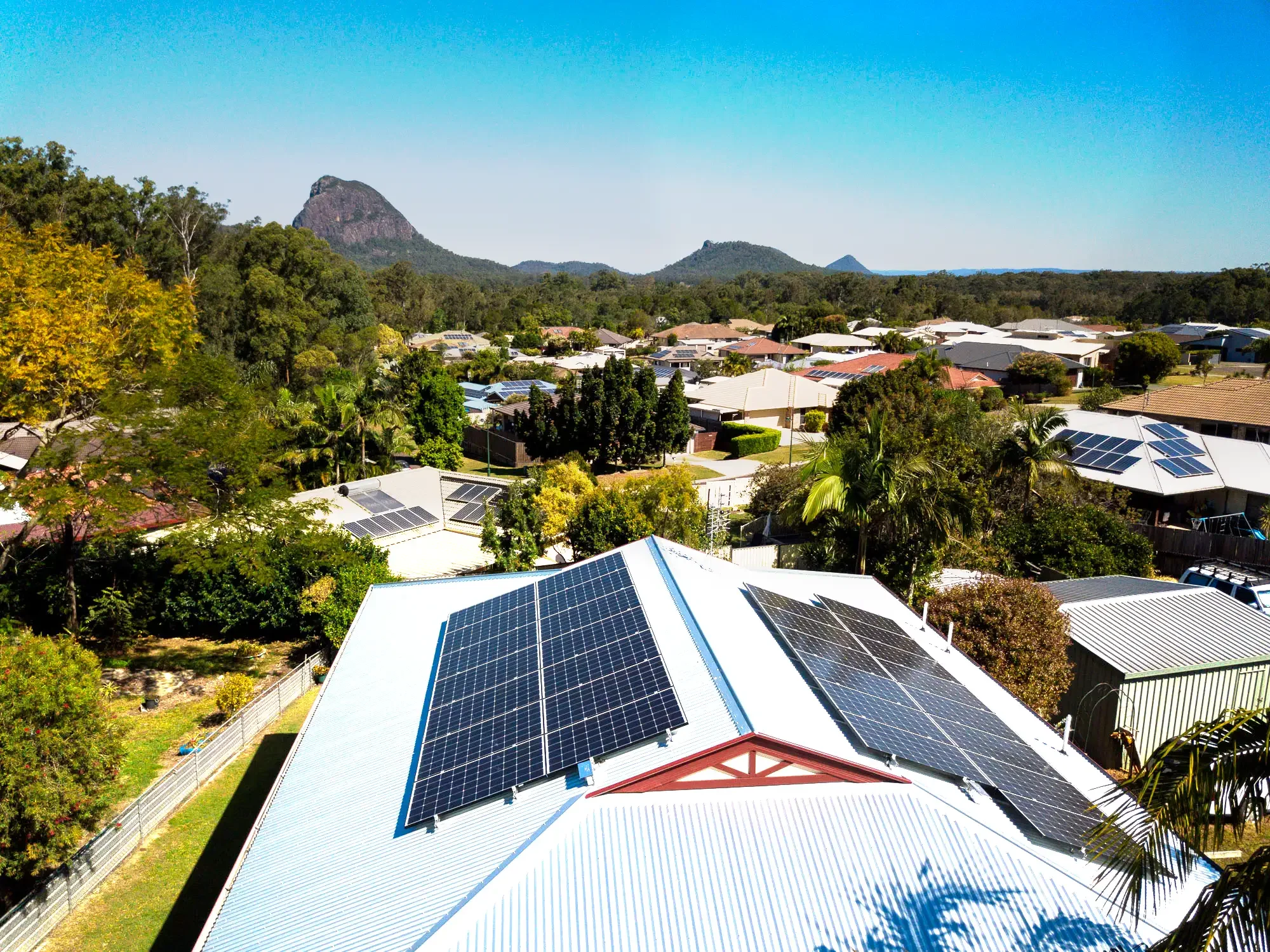 A bird's eye view of a suburban neighborhood with rooftops equipped with solar panels, surrounded by lush trees and mountains in the background under a clear blue sky.
