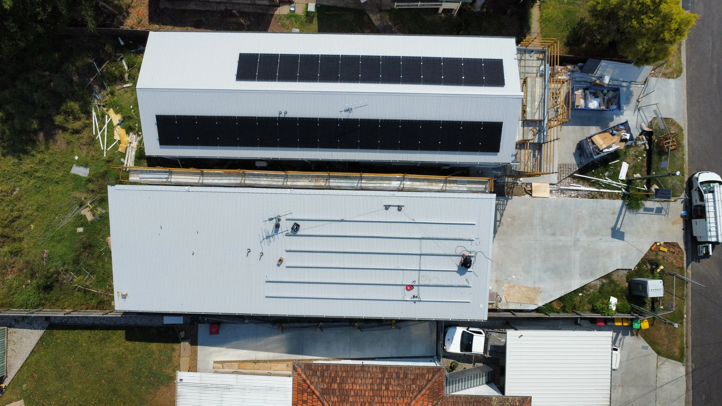 Aerial view of a construction site on a building with a white roof. Solar panels are installed on the top section, and various construction materials and equipment are scattered on the roof and surrounding area. A small highway and some greenery are 