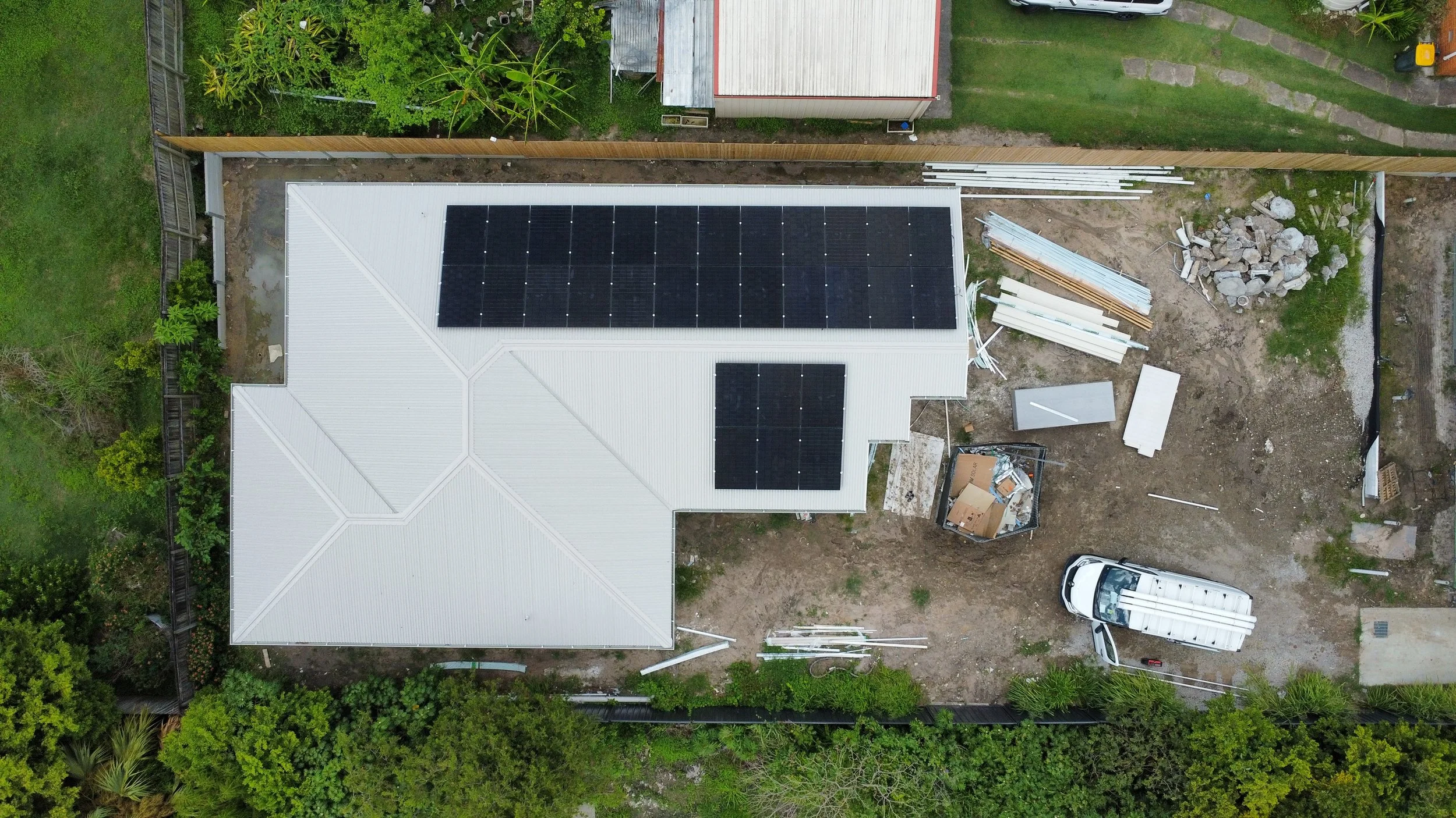 Aerial view of a house with a white roof and black solar panels, construction materials and equipment around, a white van parked on a dirt area, green trees and fenced yard.