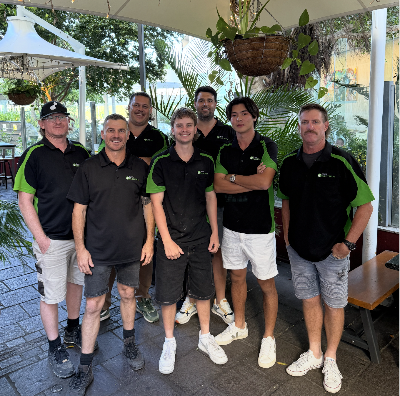 Group of seven men standing together at an outdoor cafe or patio, wearing black and green uniforms with a company logo, surrounded by plants and hanging baskets, smiling at the camera.