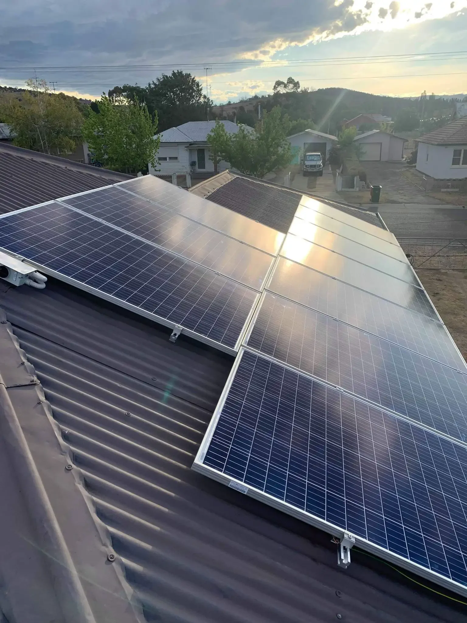 Solar panels installed on a residential roof with a neighborhood scene in the background during sunset.