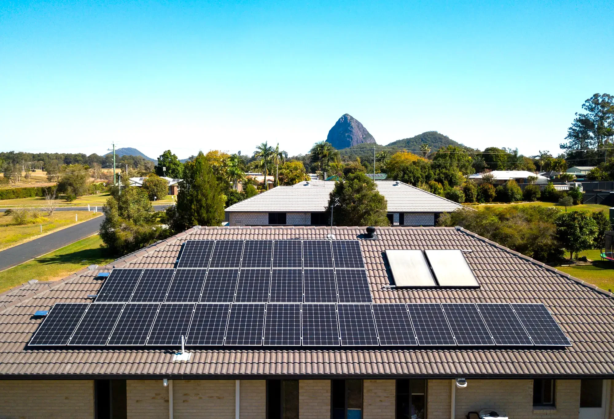 A house with solar panels on the roof, with trees and other houses in the background, and a mountain in the distance.