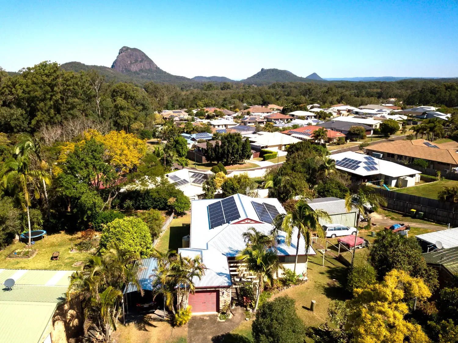 Aerial view of a neighborhood with houses that have solar panels on roofs, surrounded by lush green trees and a distant mountain range under a clear blue sky.