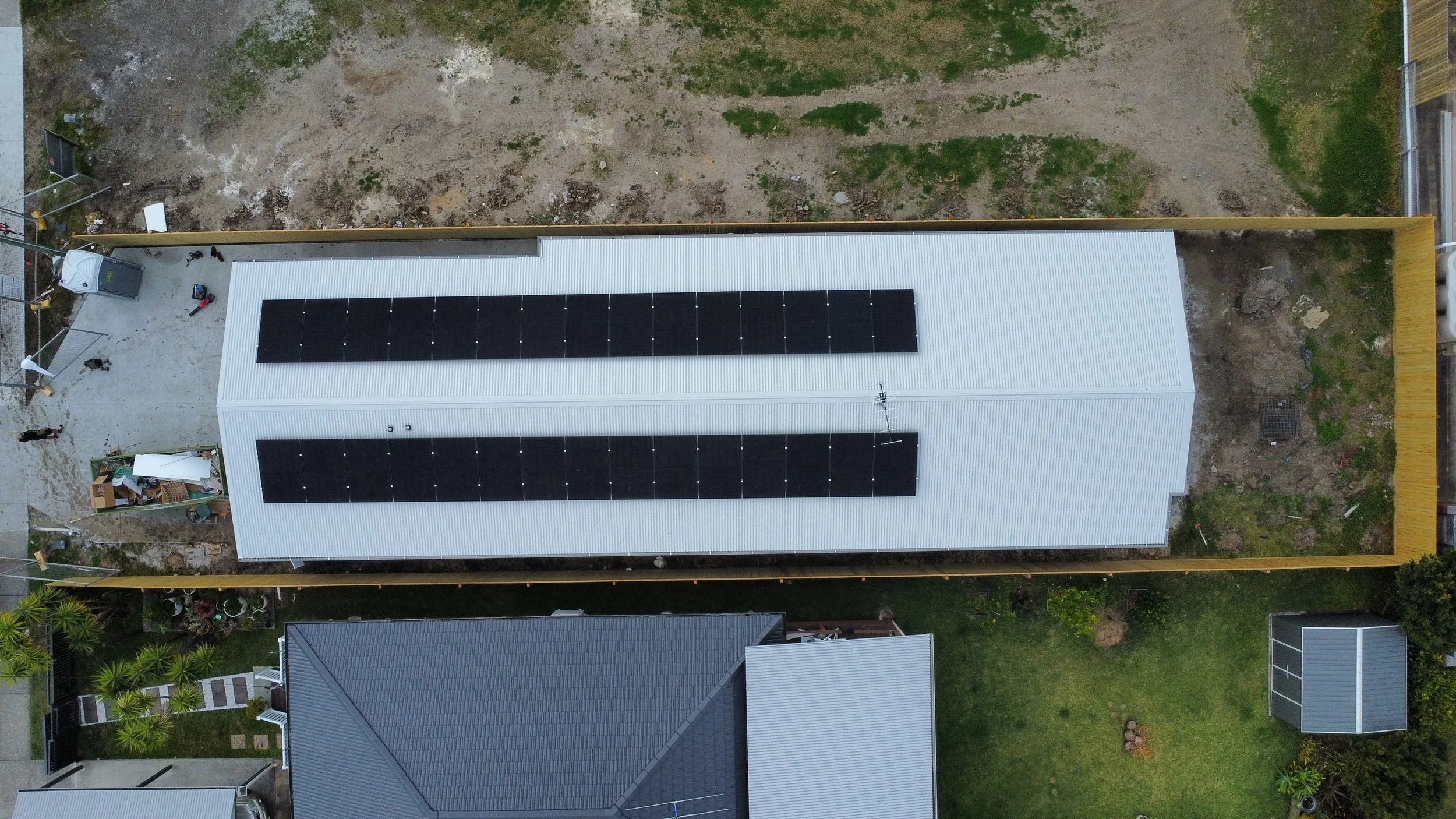 Aerial view of a residential backyard with a metal shed that has solar panels installed on its roof, surrounded by a wooden fence and neighboring houses.