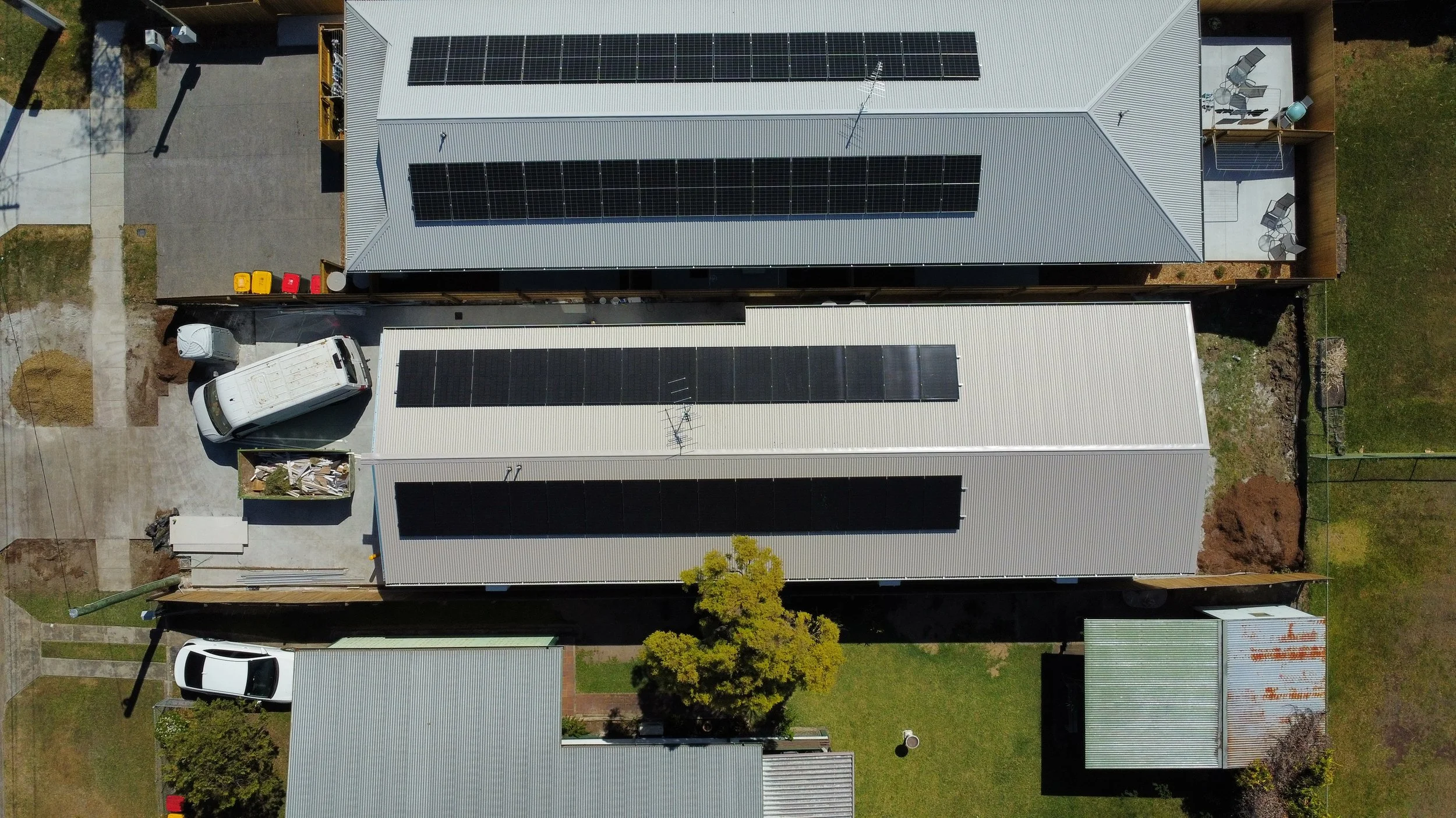 An aerial view of multiple buildings with solar panels on their roofs, a parked white utility van, a smaller building adjacent to a green lawn with a tree, and various outdoor furniture and utility items.