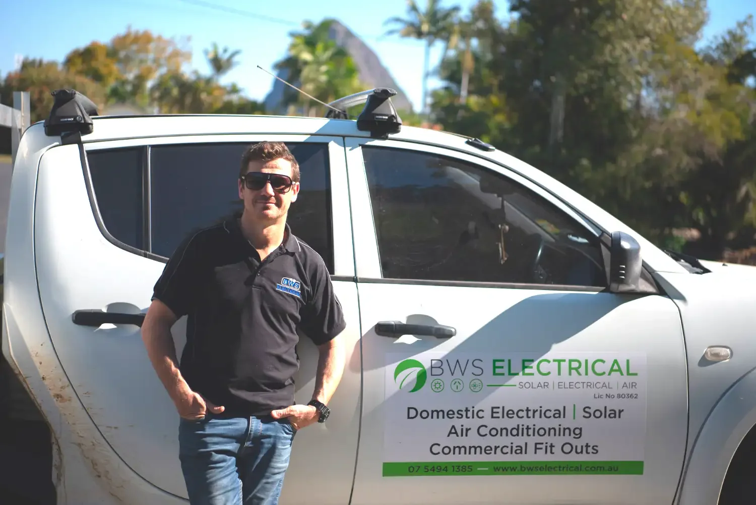 A man wearing sunglasses and a black polo shirt stands beside a white utility vehicle with company branding for BWS Electrical. The vehicle has text advertising domestic electrical, solar, air conditioning, and commercial fit-outs, along with contact