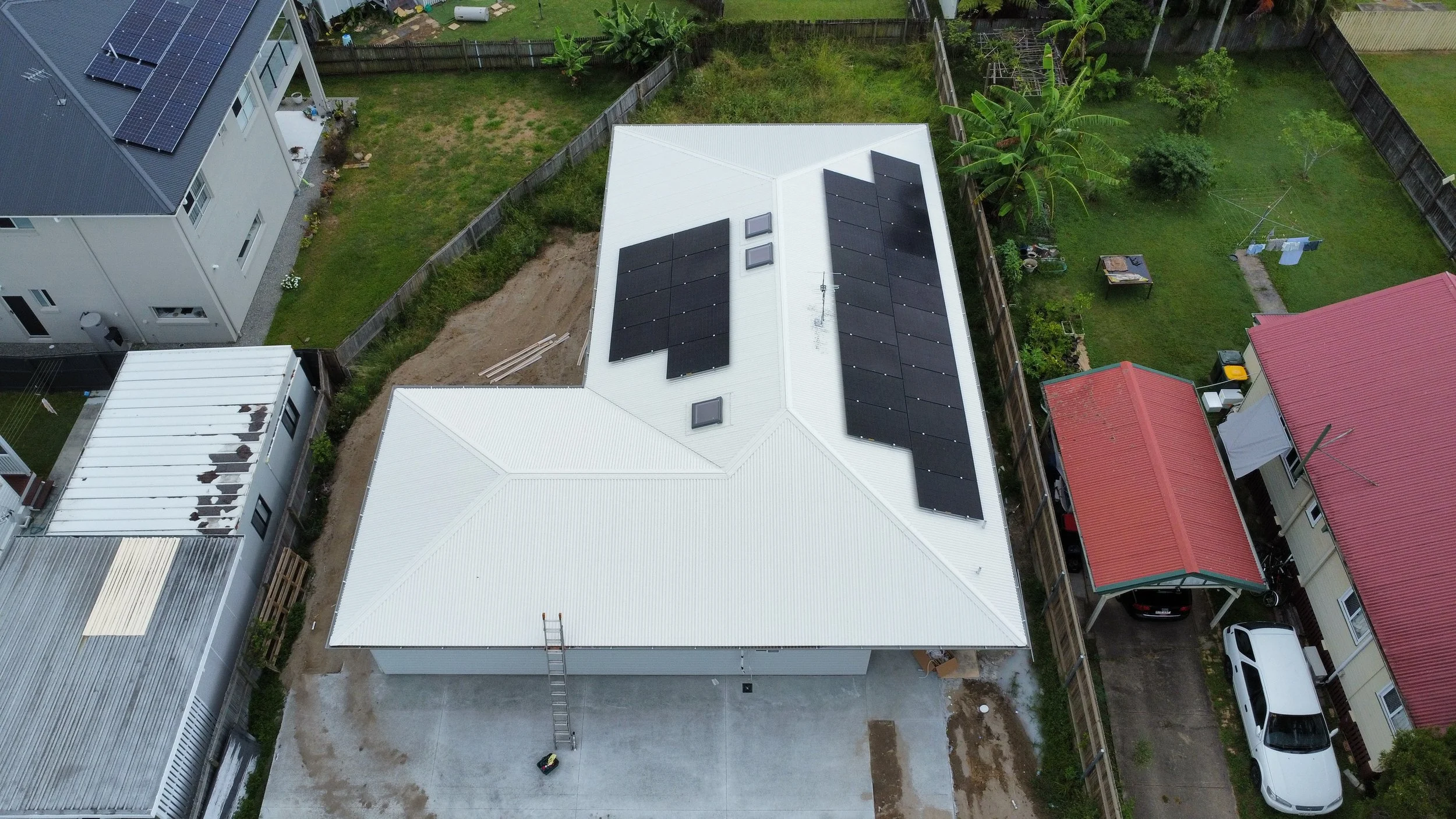 Aerial view of a white house with solar panels on the roof, next to neighboring houses with different colored roofs, surrounded by fences, yard, and parked vehicles.