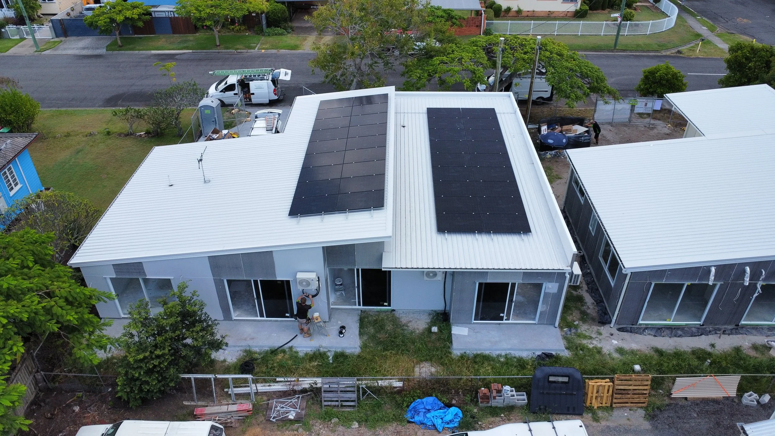 Aerial view of a residential house with solar panels on its roof, a worker installing or repairing an air conditioning unit outside, neighboring houses, trees, vehicles, and construction materials in the yard.