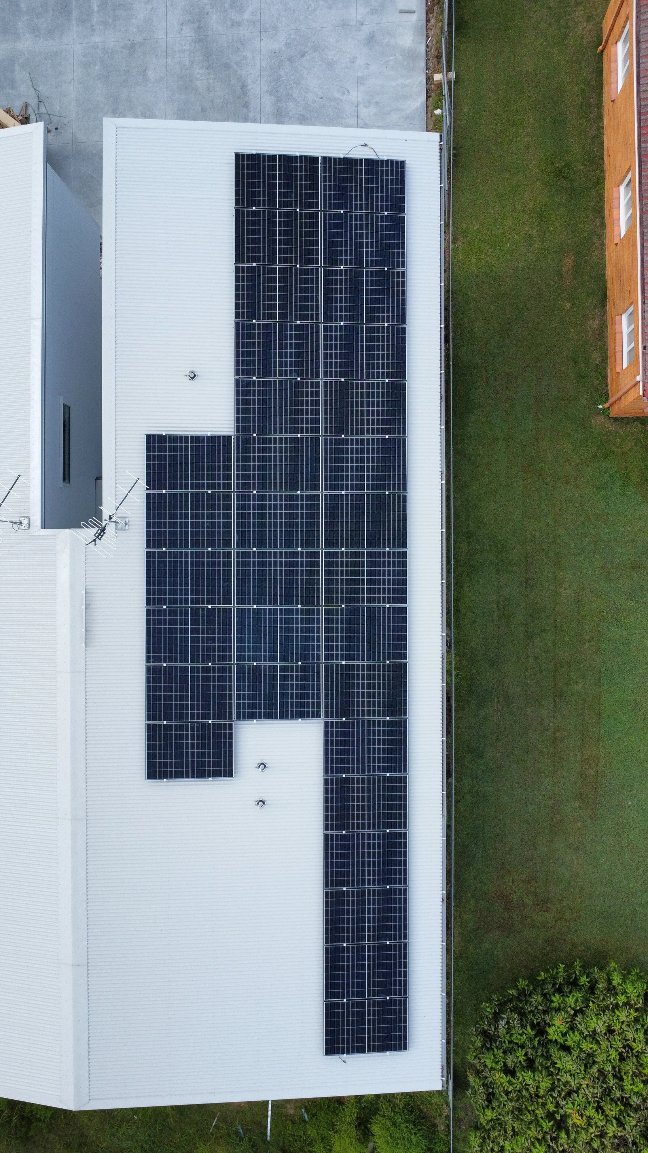 Aerial view of a white metal roof with multiple solar panels installed, next to a grassy yard and neighboring buildings.
