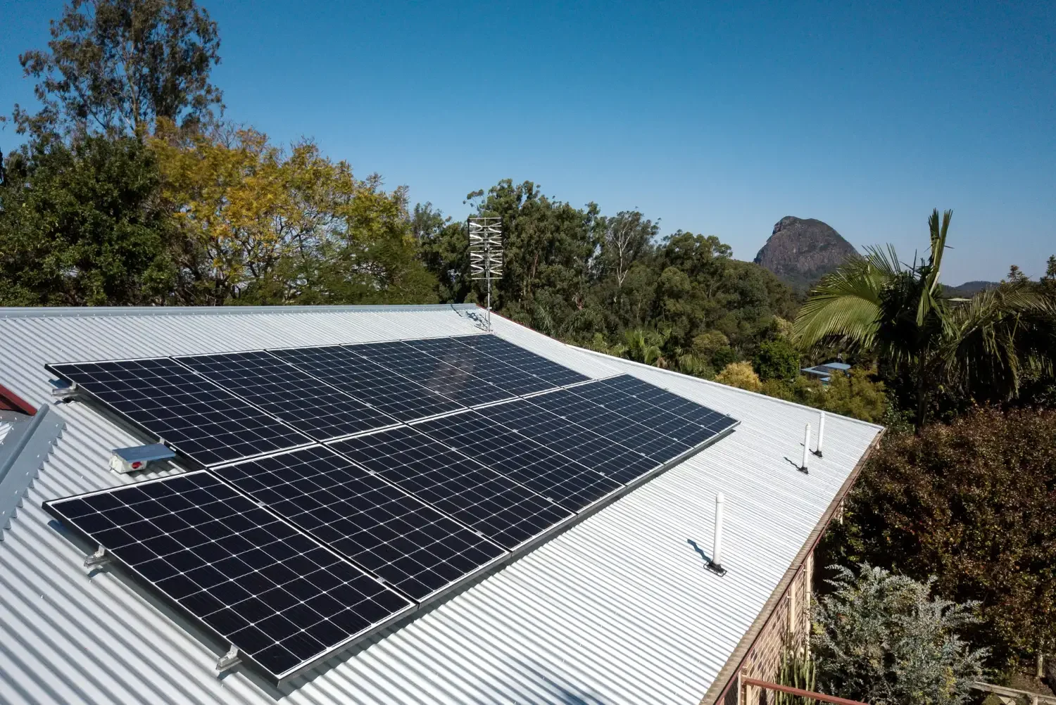 Solar panels installed on a corrugated metal roof surrounded by trees and a mountain in the distance under a blue sky.