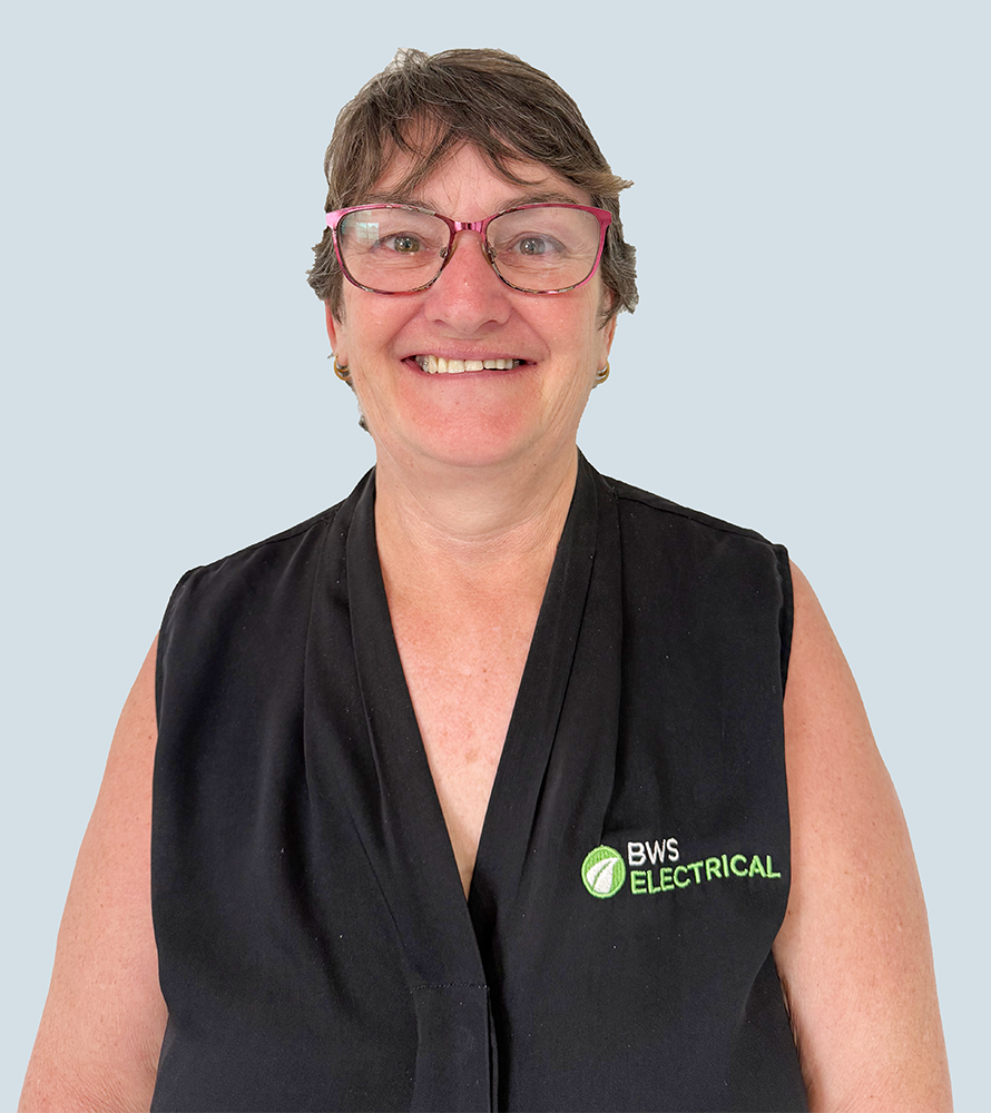 A smiling woman with short gray hair, wearing pink glasses and a black sleeveless shirt with the logo and text "BWS Electrical" embroidered on it, standing against a light blue background.