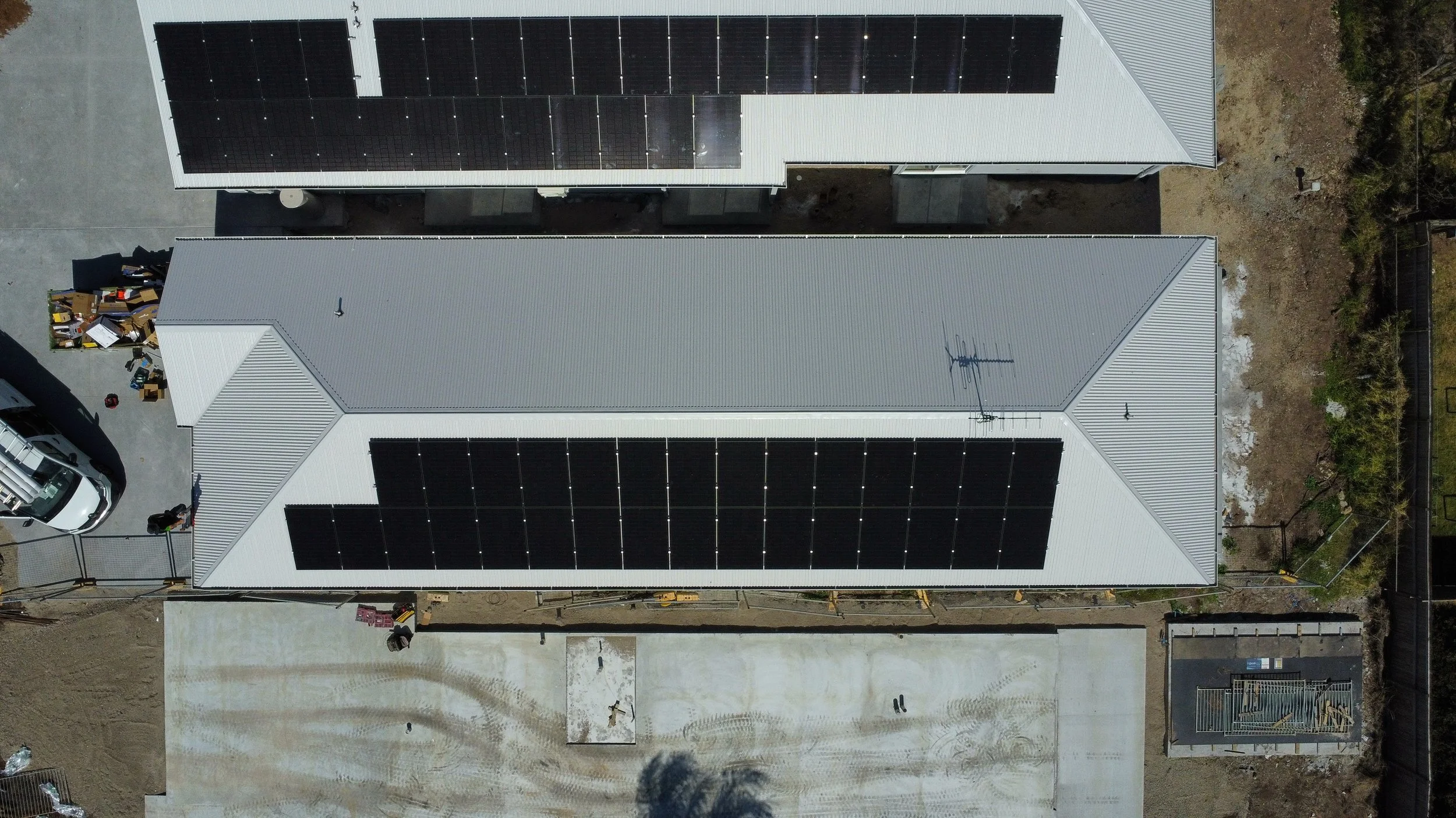 Aerial view of a building with solar panels on its white metal roof, showing a grey area under construction in front and surrounding landscape with trees and fencing.