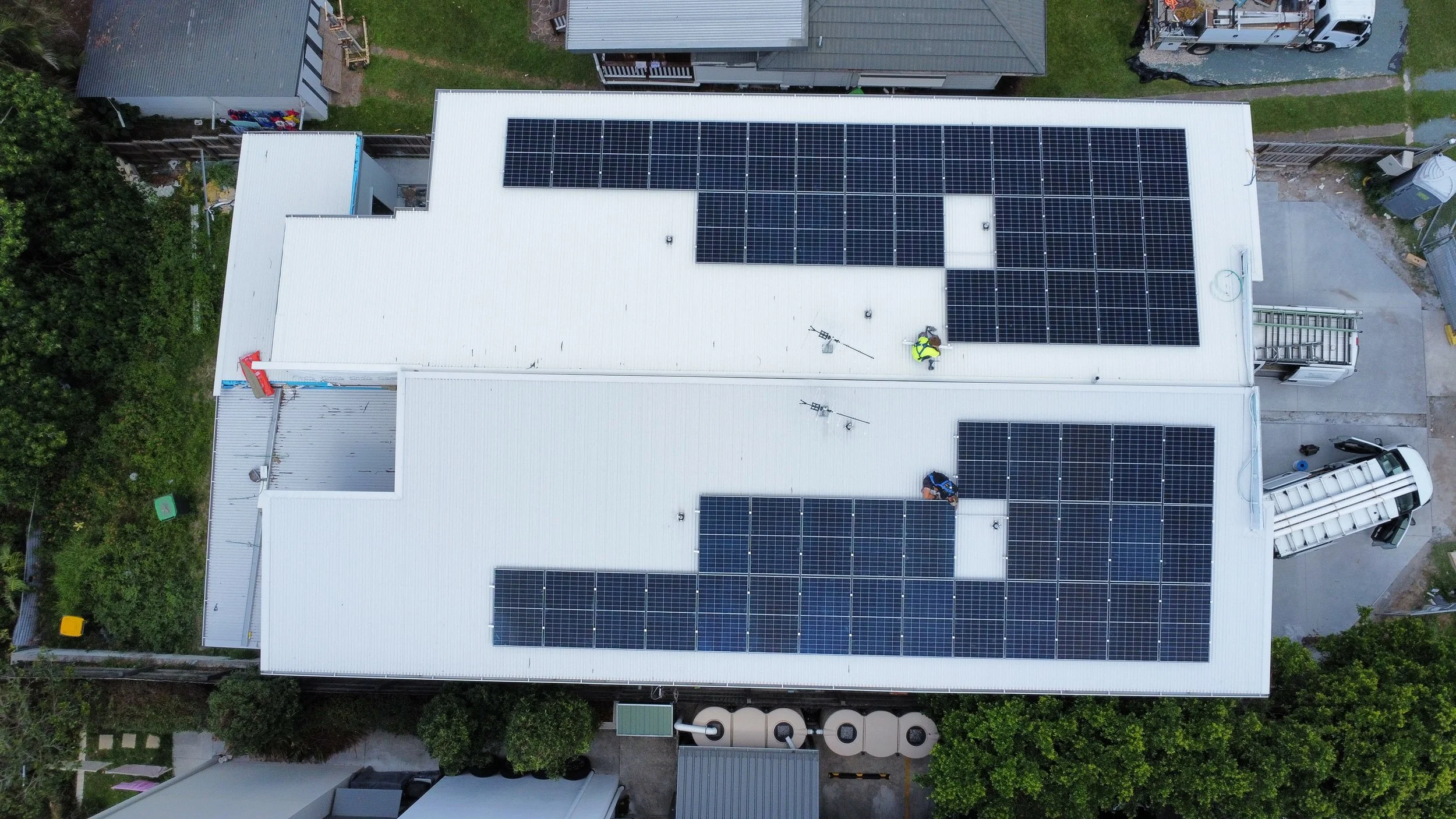 Aerial view of a building with multiple solar panels installed on its white metal roof, with trees and neighboring houses visible around.