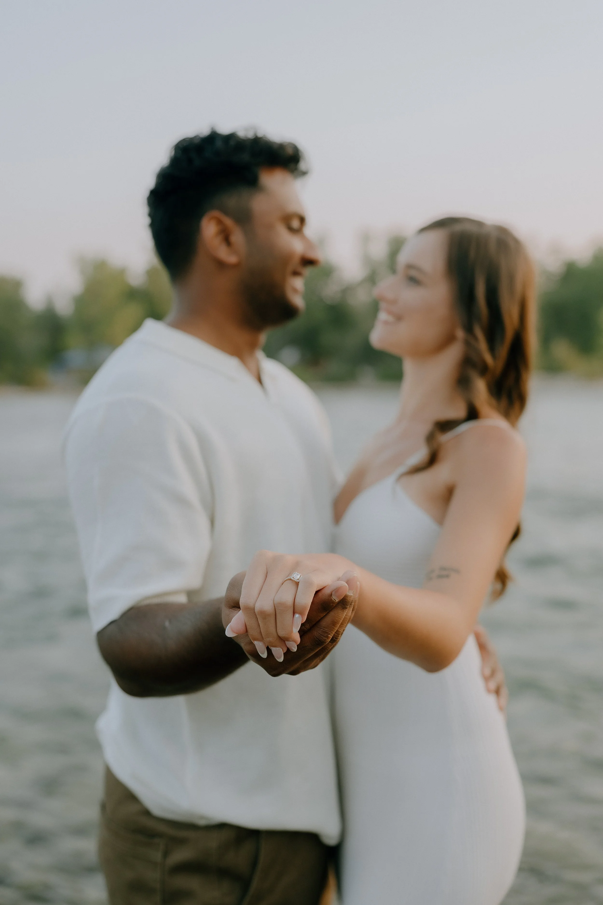 A couple holds hands and faces each other affectionately by the water, with trees in the background.