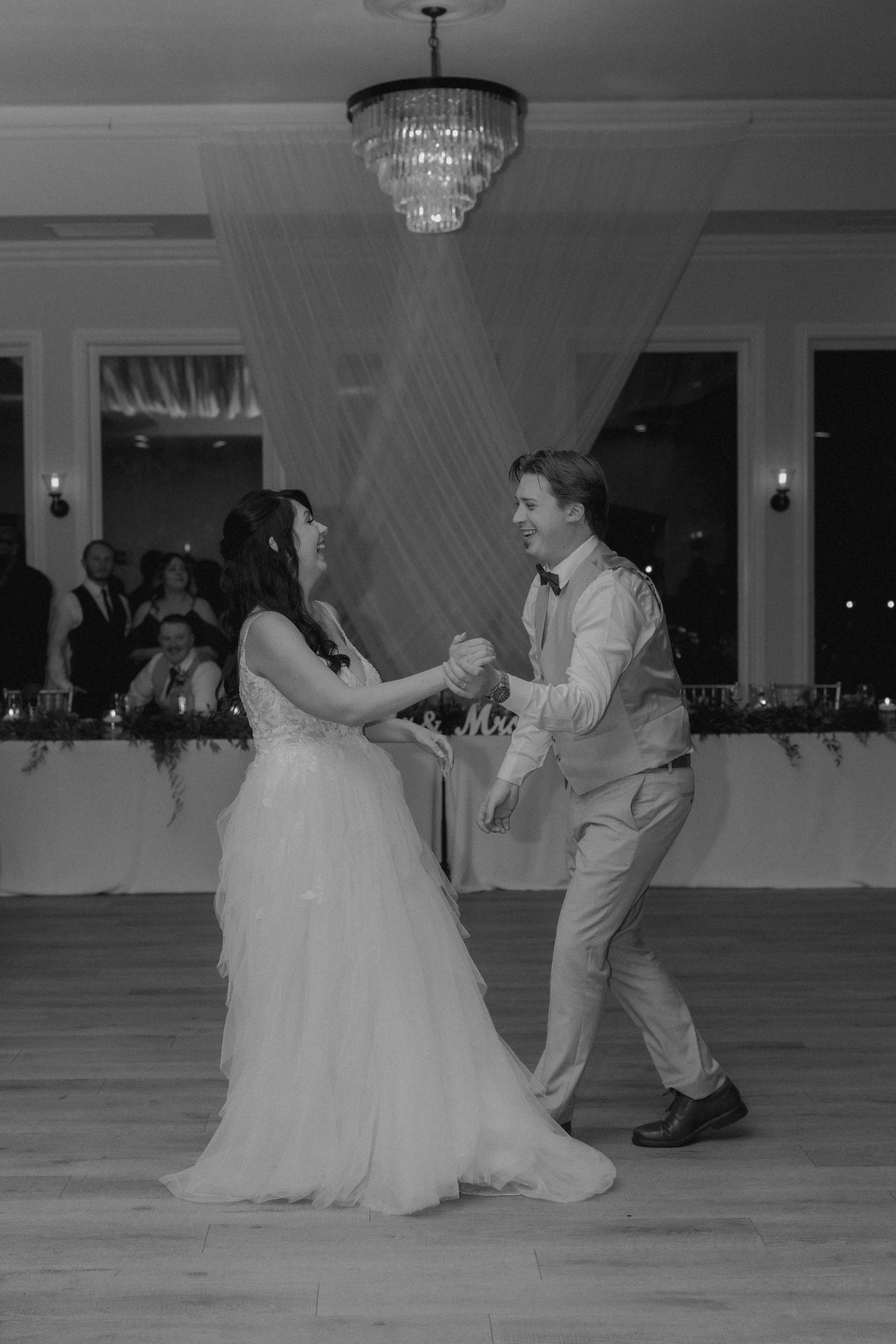 A bride and groom dancing together at their wedding reception, smiling and holding hands, with guests in the background.