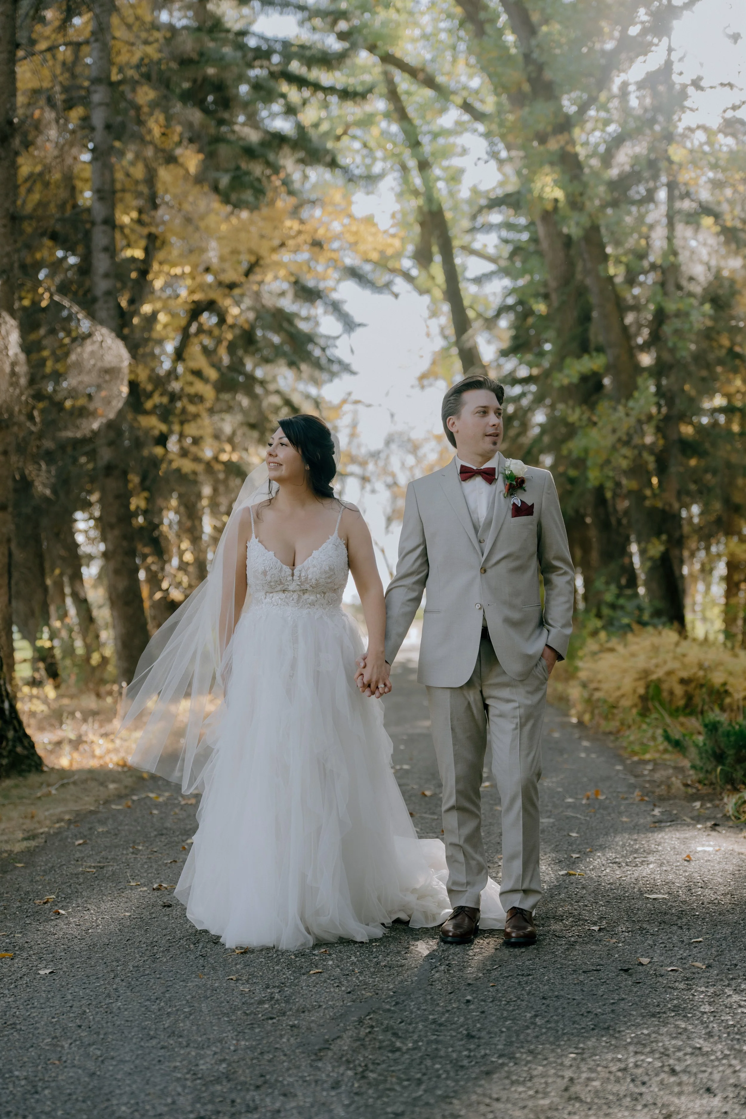 A bride and groom holding hands and walking outdoors on a trail in a wooded area during fall, with trees and foliage around them.