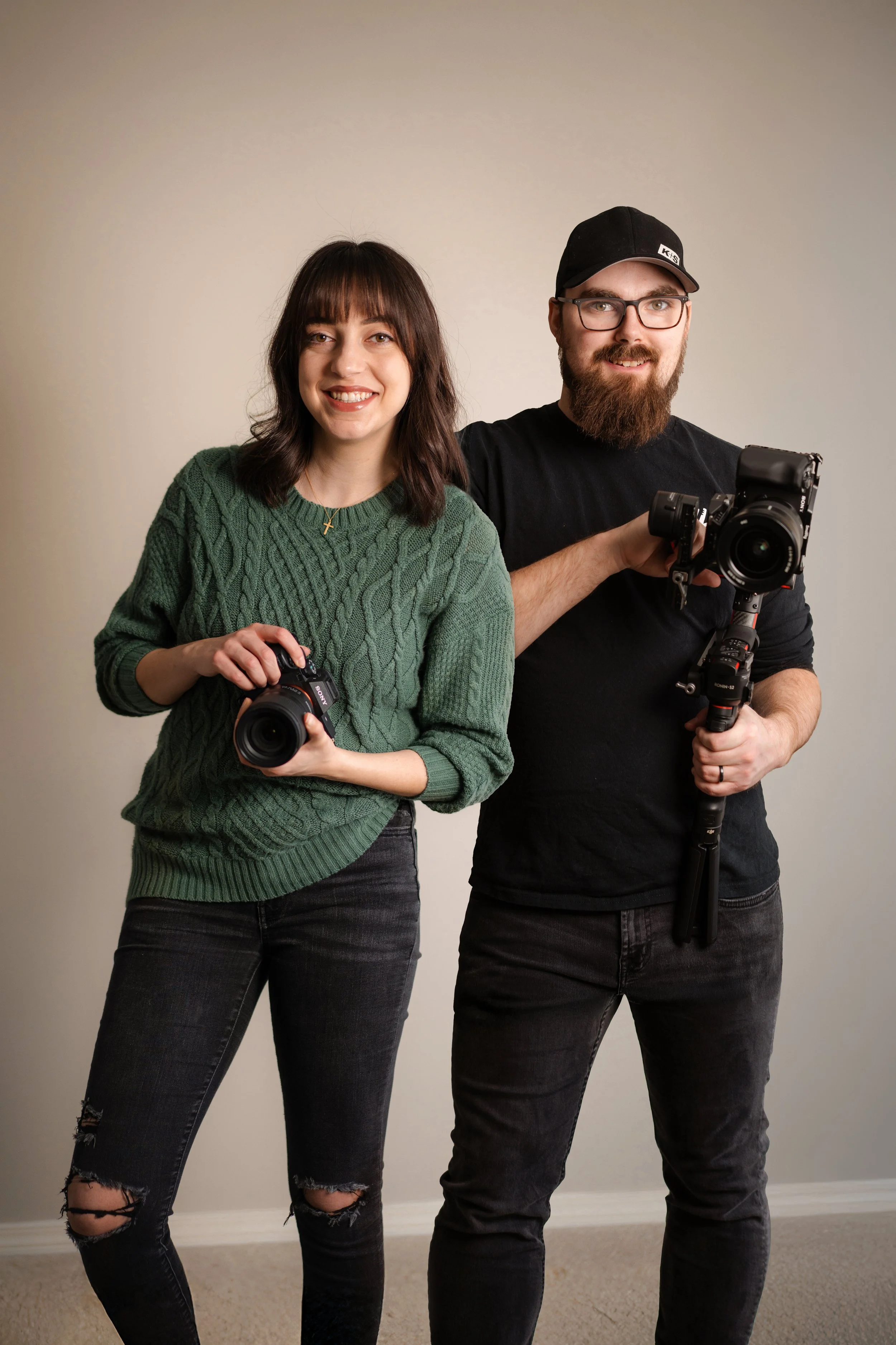 A woman with dark brown hair wearing a green sweater and ripped black jeans holding a camera, standing next to a man with a beard, glasses, black cap, black shirt, and black jeans, holding a camera on a stabilizer, against a plain wall.