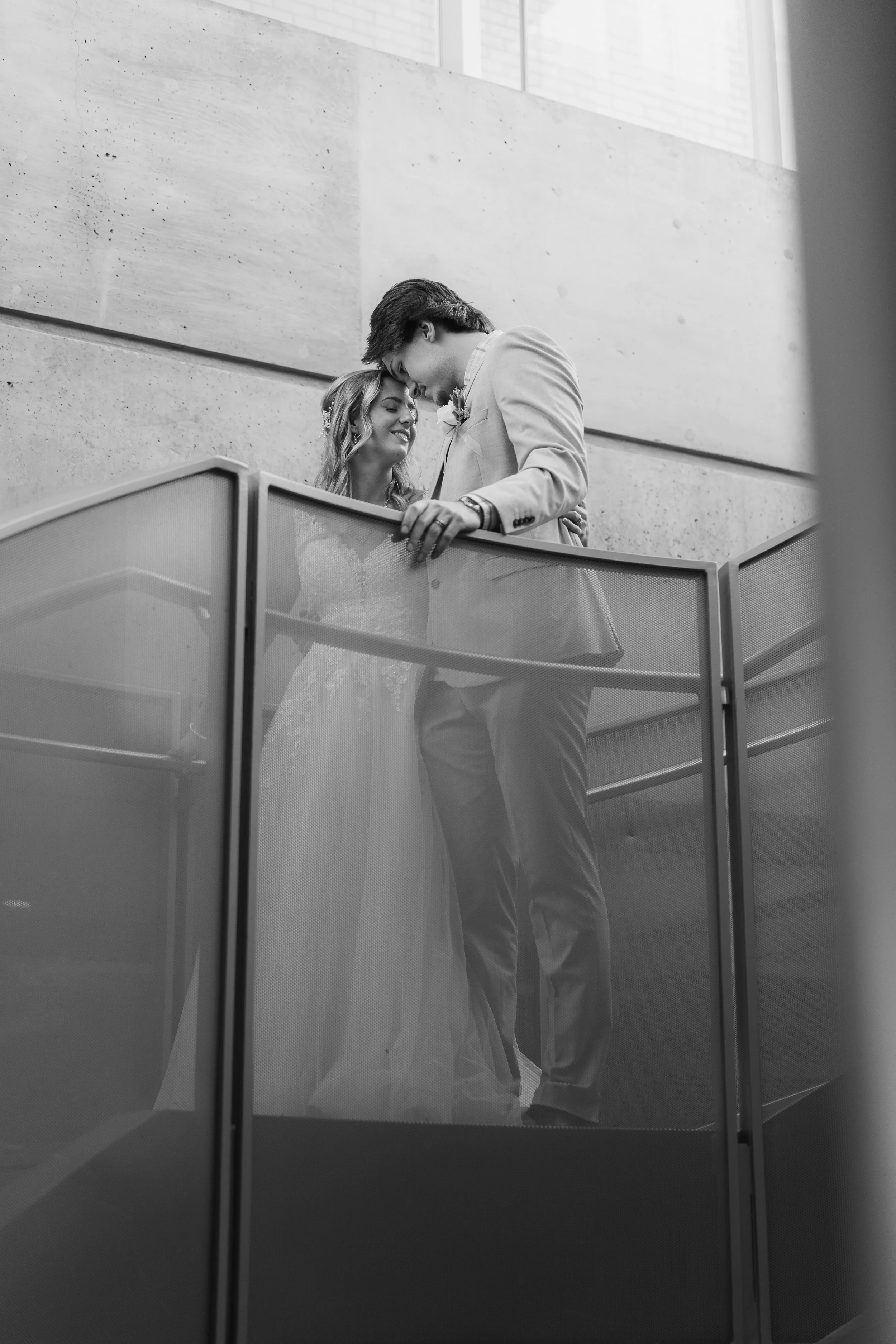 Black and white photo of a bride and groom embracing on a staircase, smiling at each other.