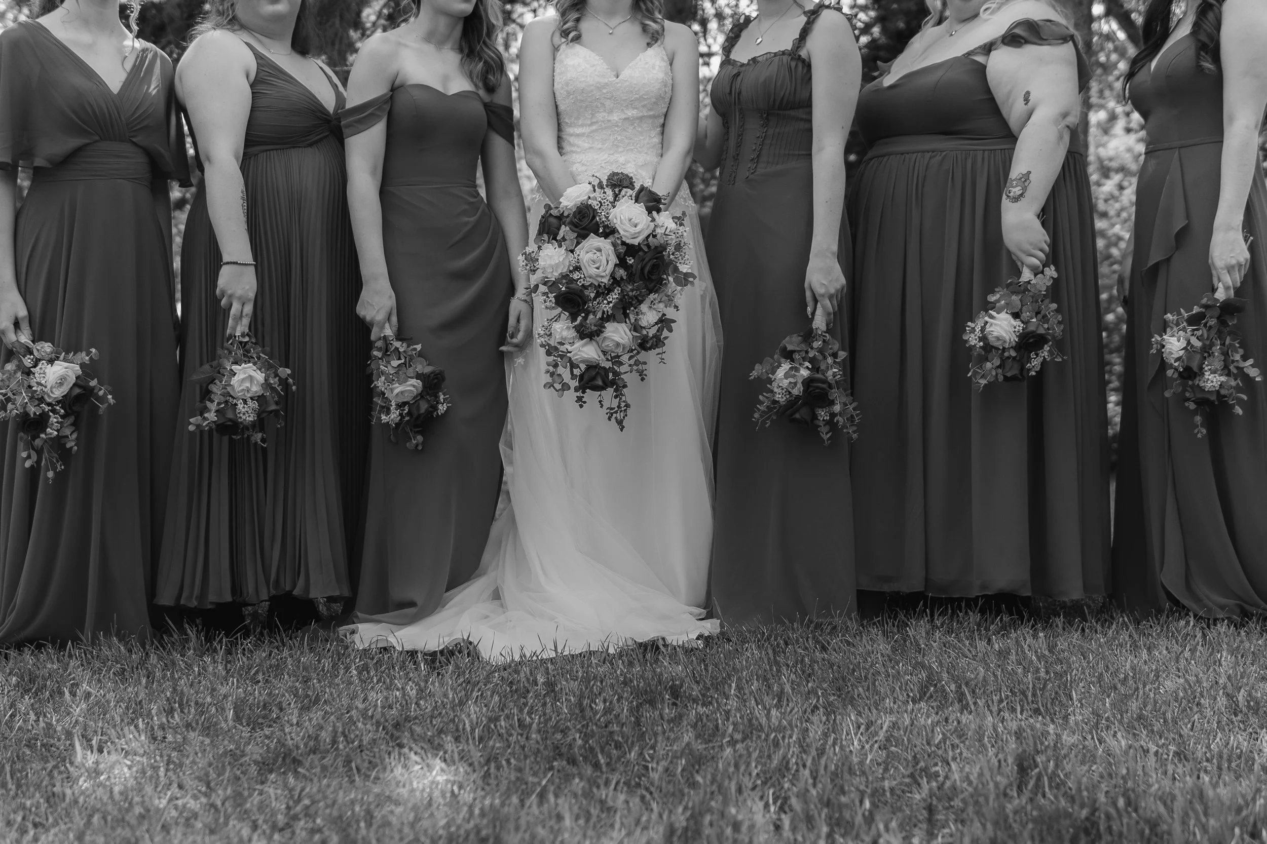 A bride in a white wedding dress holding a bouquet standing with bridesmaids in dark dresses holding bouquets, outdoors on grass.