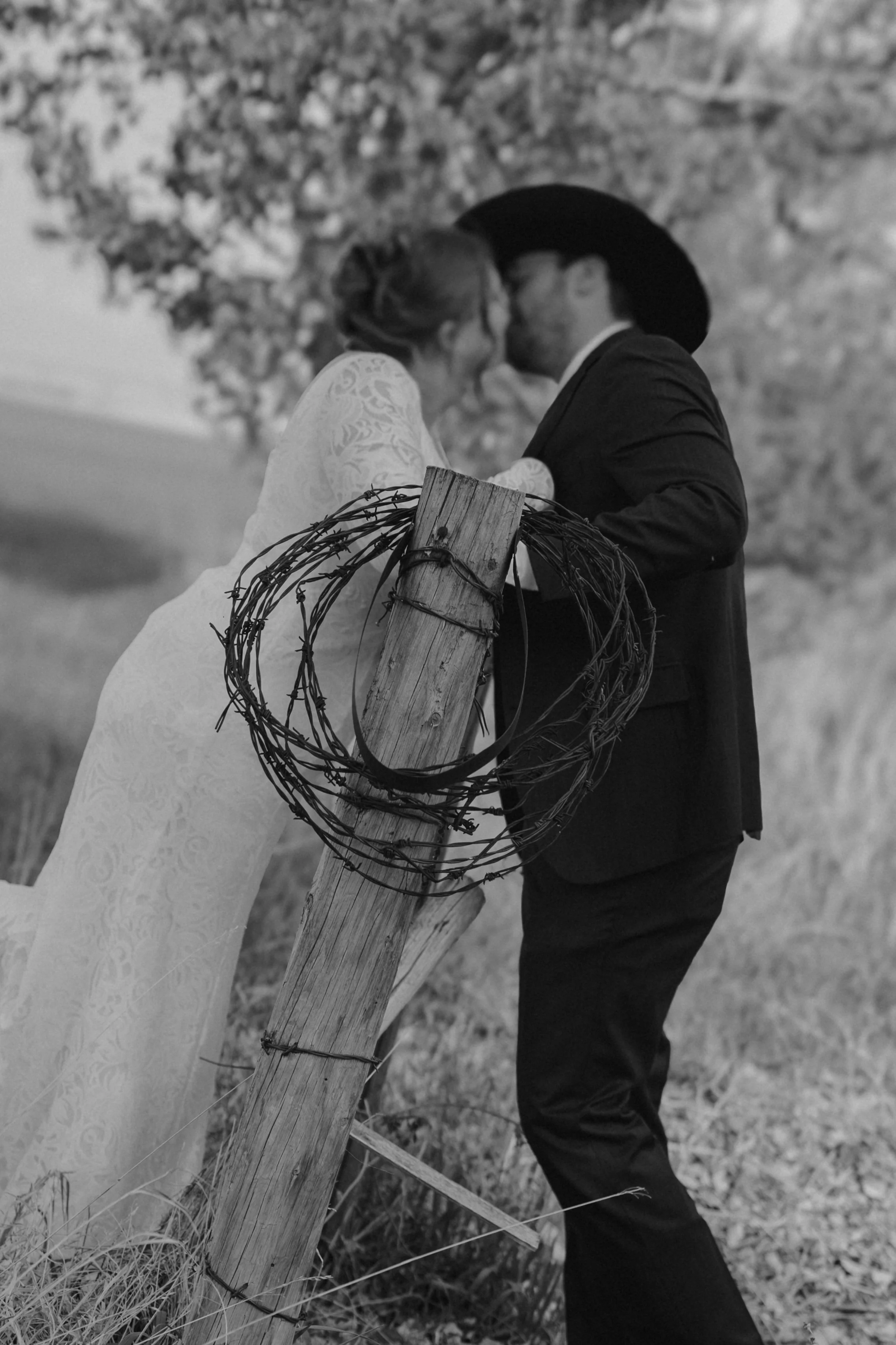 A couple sharing a kiss in a rural setting, standing behind a barbed wire fence with a wooden post.