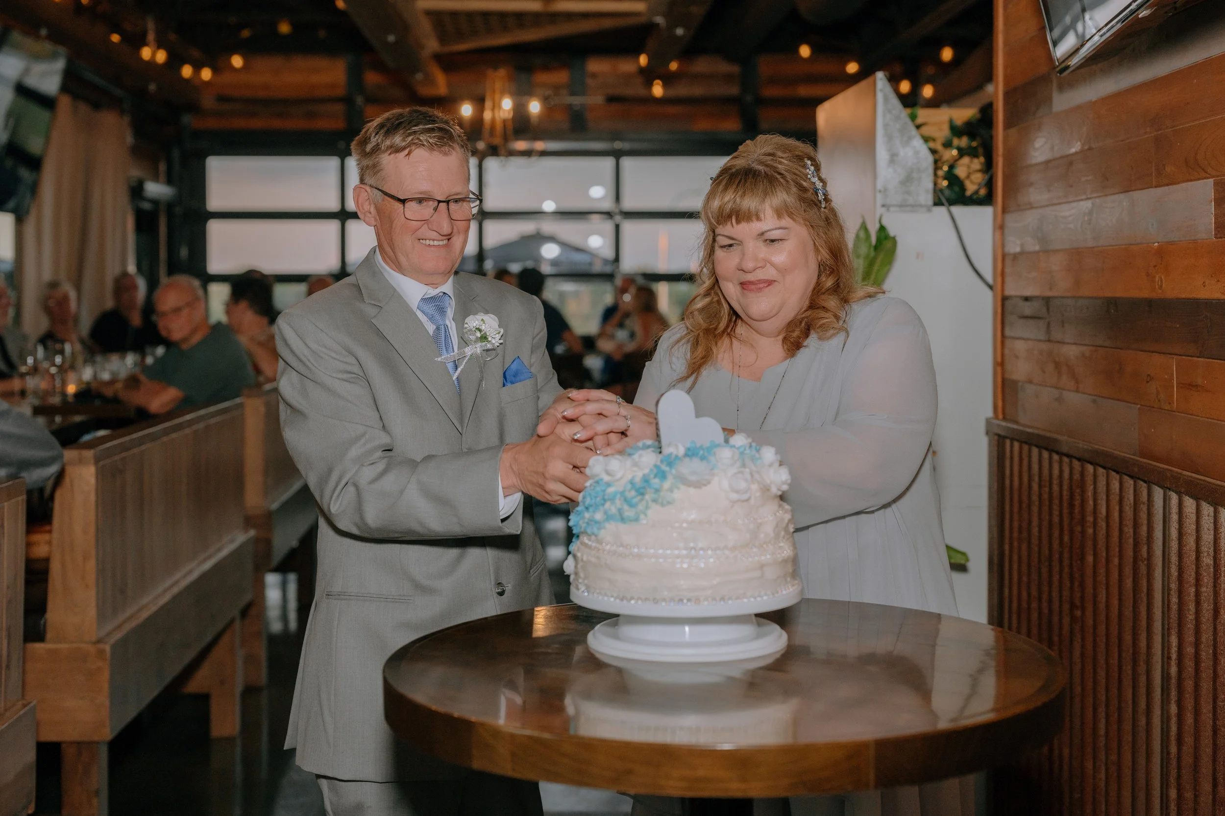 A man and woman in formal attire cutting a wedding cake together at a celebration in a warmly lit venue with other guests watching.