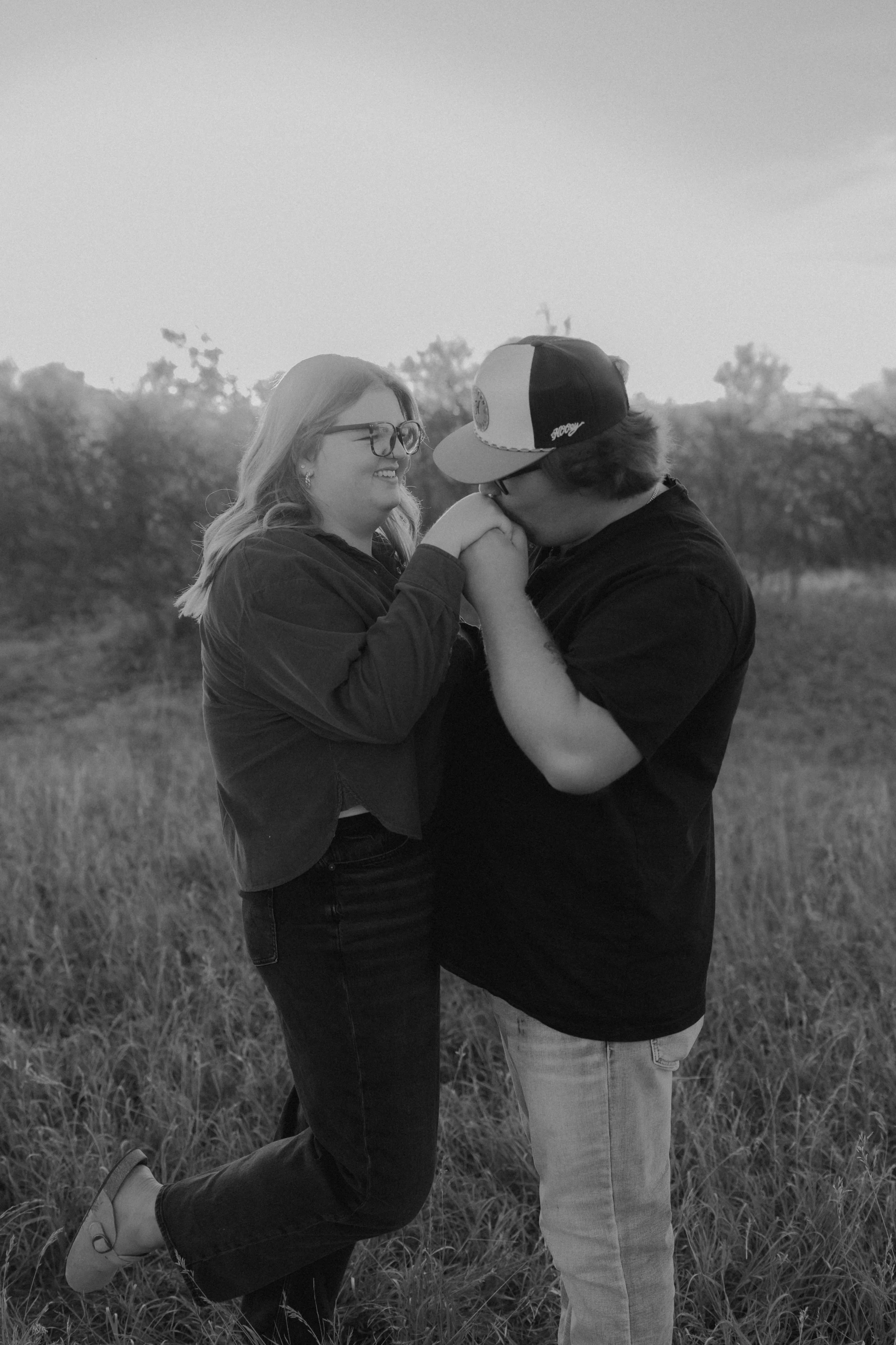 A black-and-white photo of a couple standing in a field holding hands. The woman is smiling, wearing glasses and casual clothing, and the man is kissing her hand, wearing a cap and casual clothes.