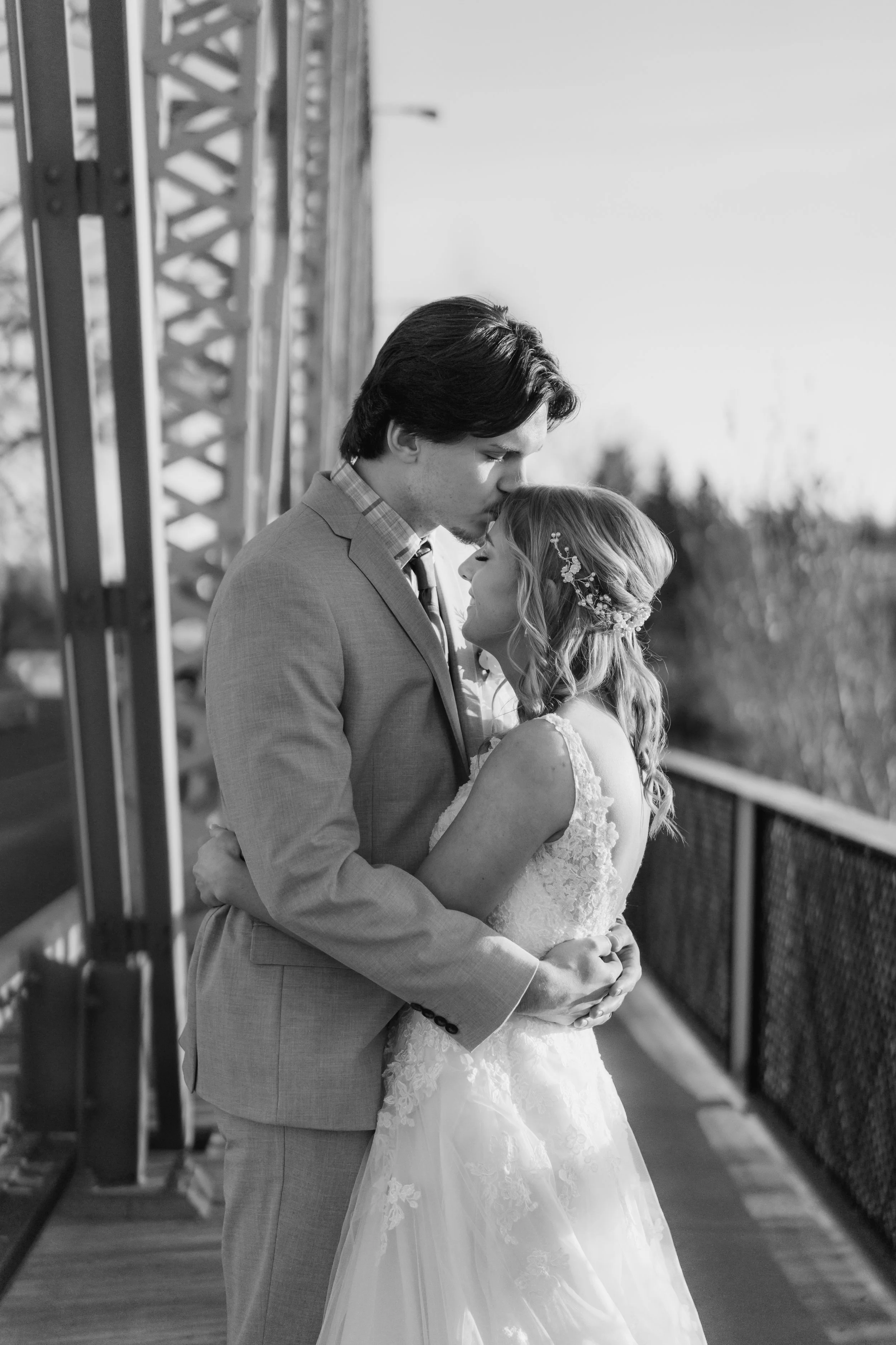 A black-and-white photo of a bride and groom kissing on a bridge during their wedding, with the groom in a suit and the bride in a lace wedding gown and floral hairpiece.