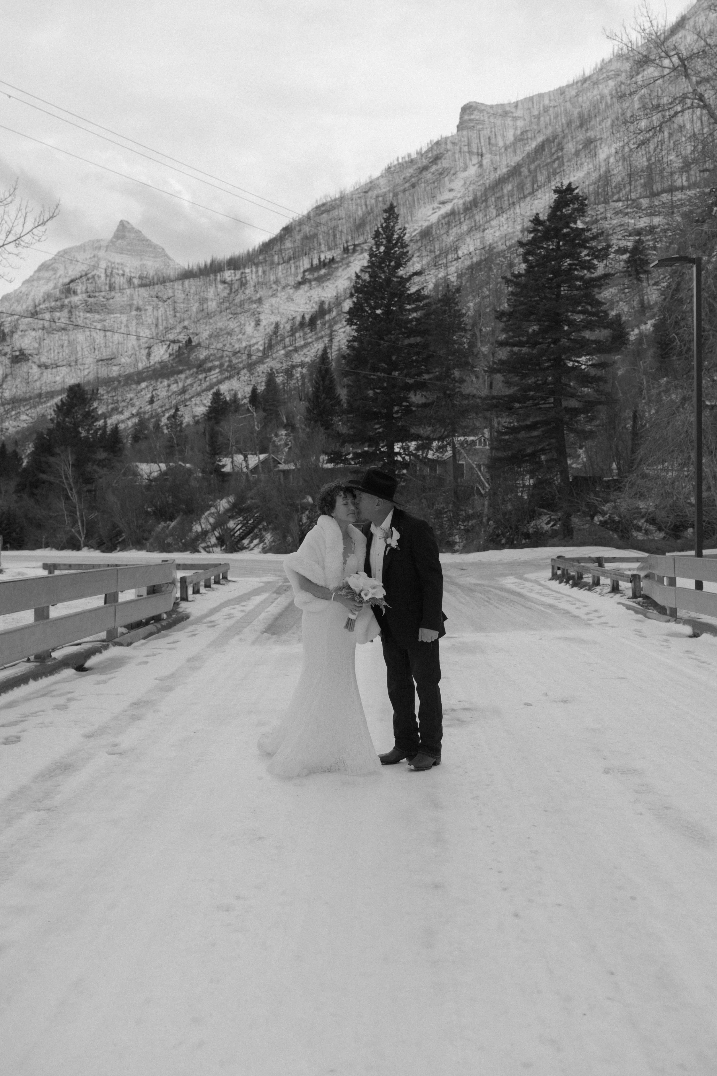 Black and white photo of a bride and groom standing on a snowy bridge, facing each other, with mountains and trees in the background.
