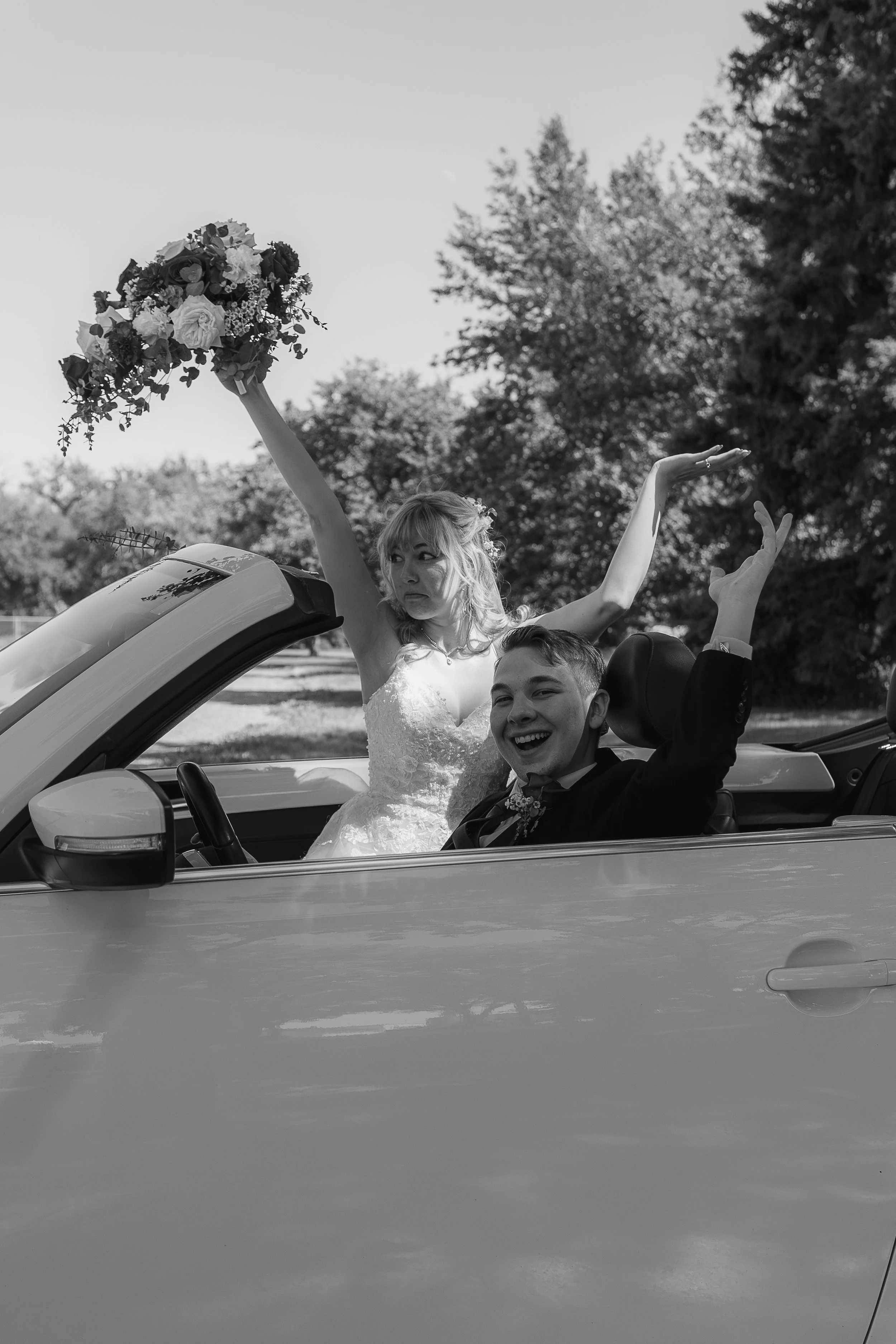 Black and white photo of a woman in a wedding dress and a man in a suit riding in a convertible car. The woman is holding a bouquet and has her hands raised, while the man is smiling and waving. Trees are visible in the background.