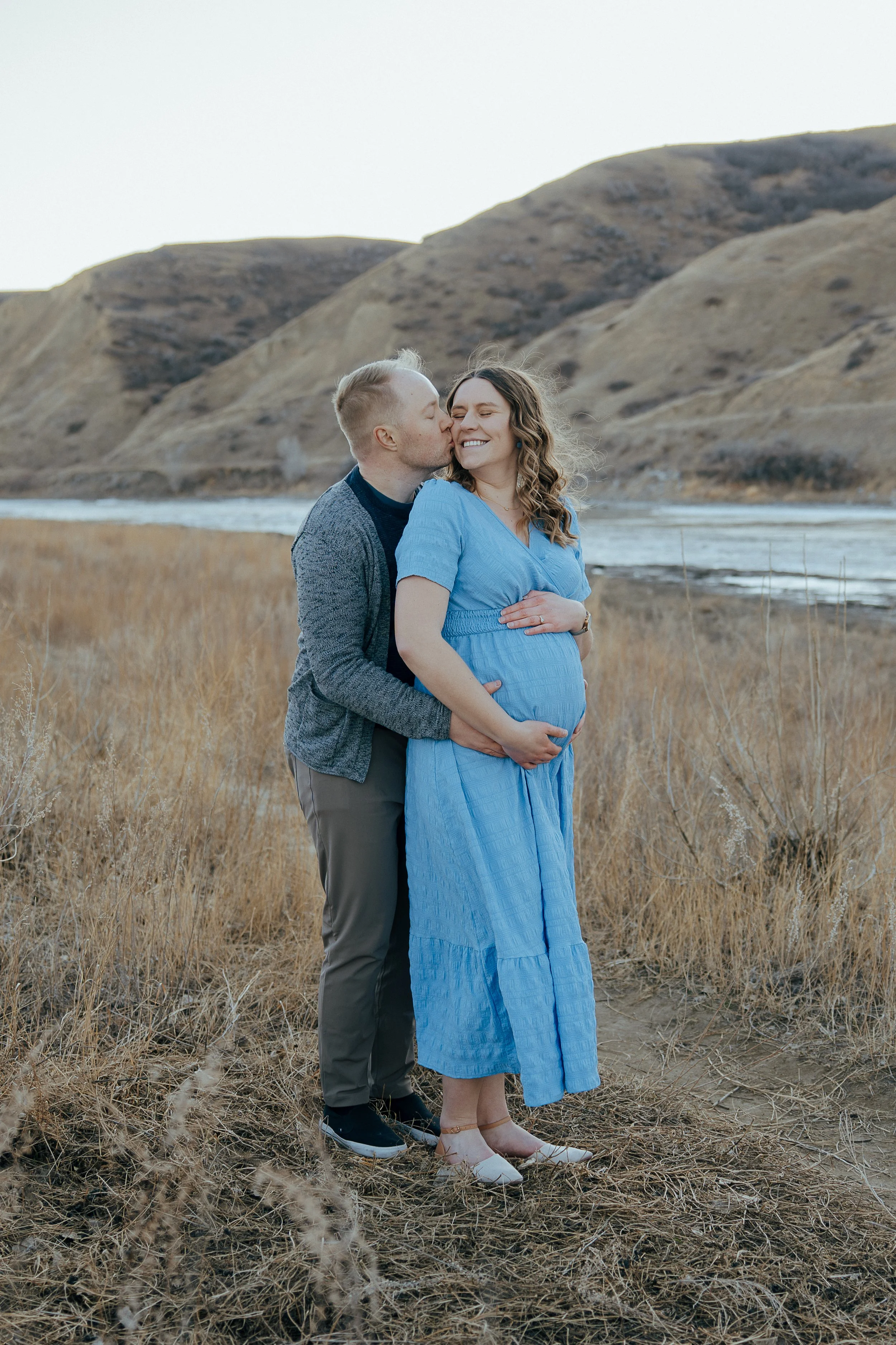A pregnant woman in a blue dress stands outdoors on grass, smiling as her partner kisses her cheek. The man is dressed casually in a gray jacket and dark pants, standing behind her with his arms around her. The landscape features a river and rolling, brown hills in the background.