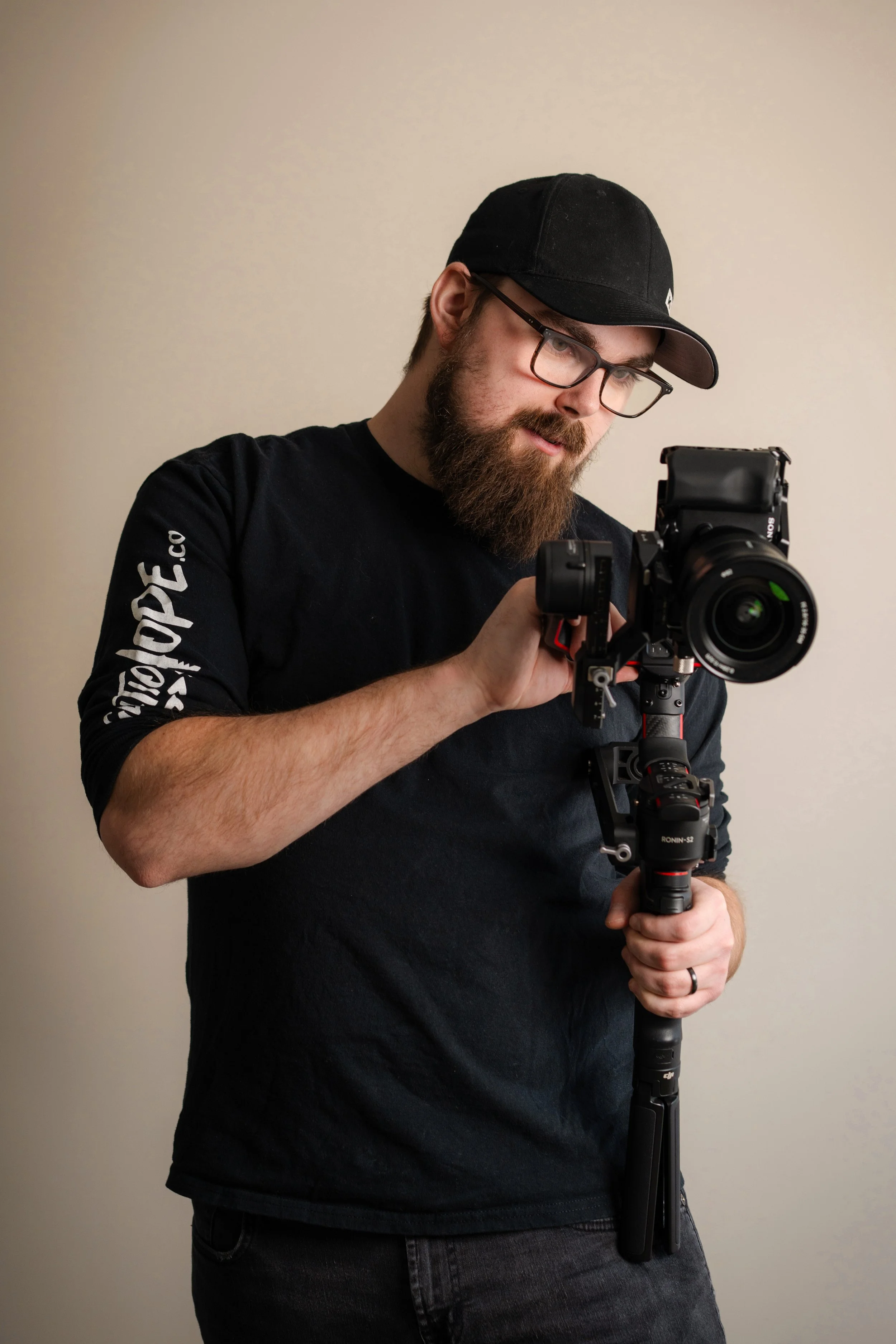 A man with a beard wearing a black baseball cap, black shirt, and glasses, holding a professional camera on a stabilizer, looking at the camera's screen.