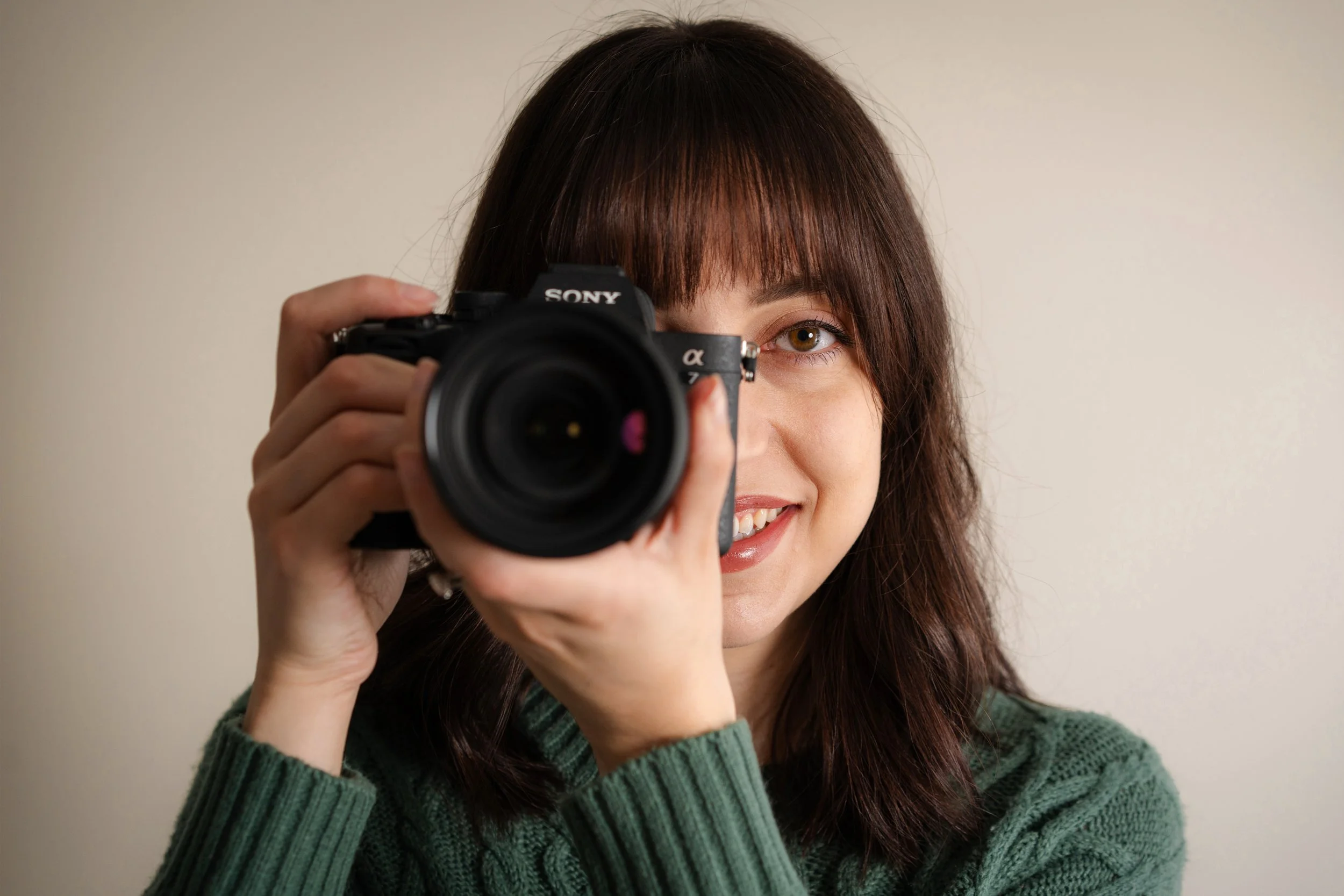 A woman with brown hair and green eyes takes a photo with a black Sony camera, smiling at the camera.
