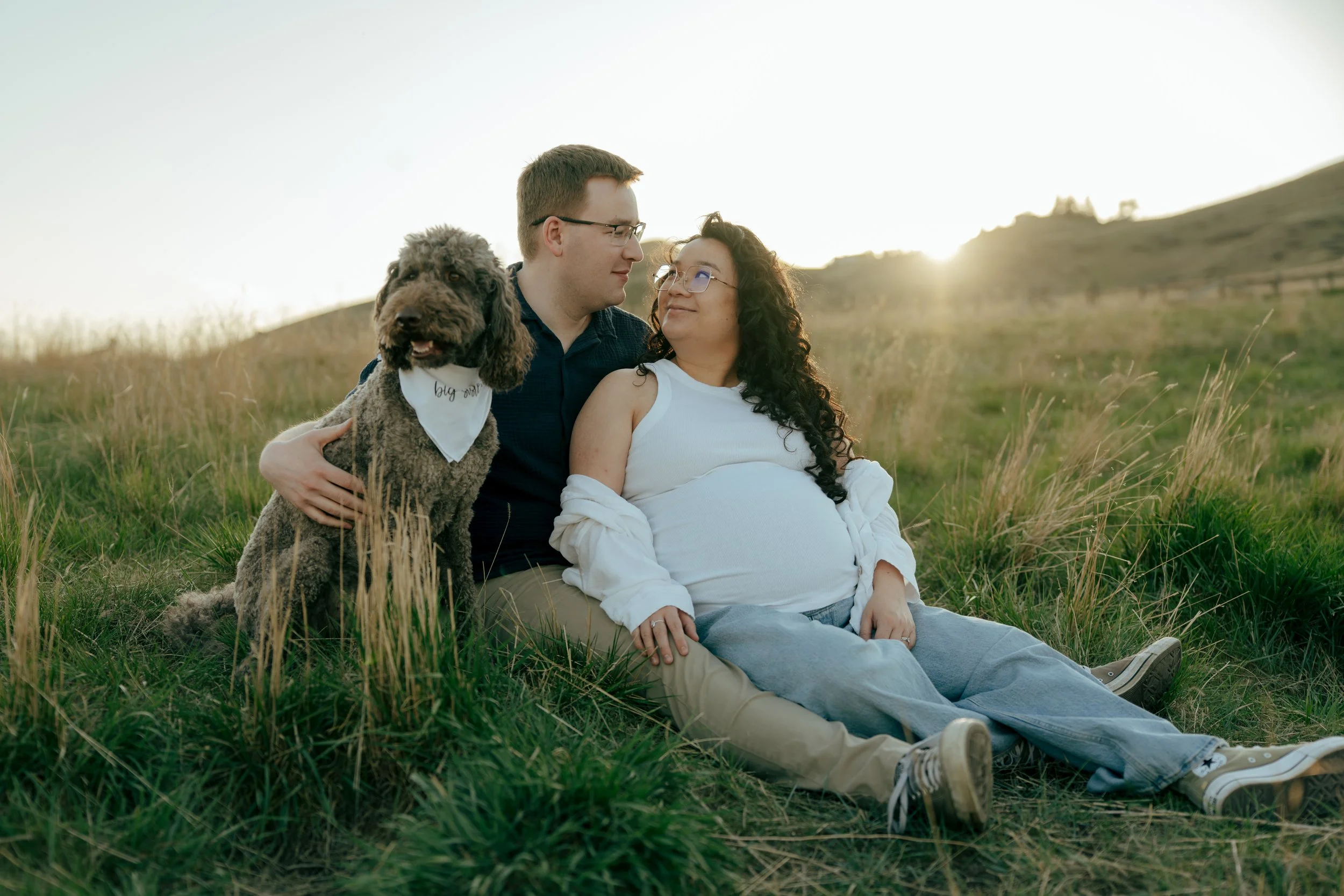 A couple sitting on grass in a field during sunset with their dog, looking lovingly at each other.