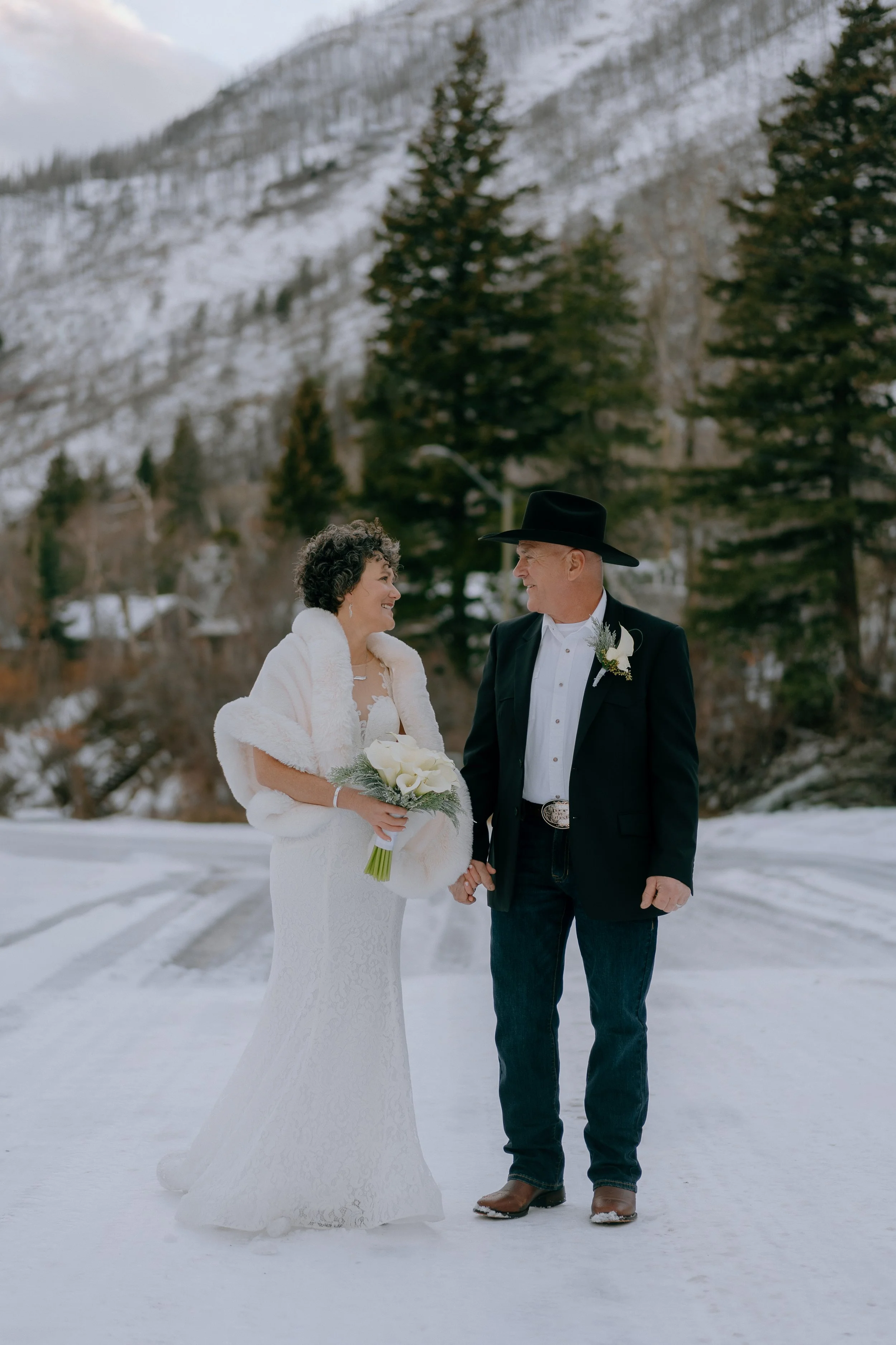 A bride and groom holding hands and smiling at each other outdoors in a snowy landscape with trees and mountains in the background.