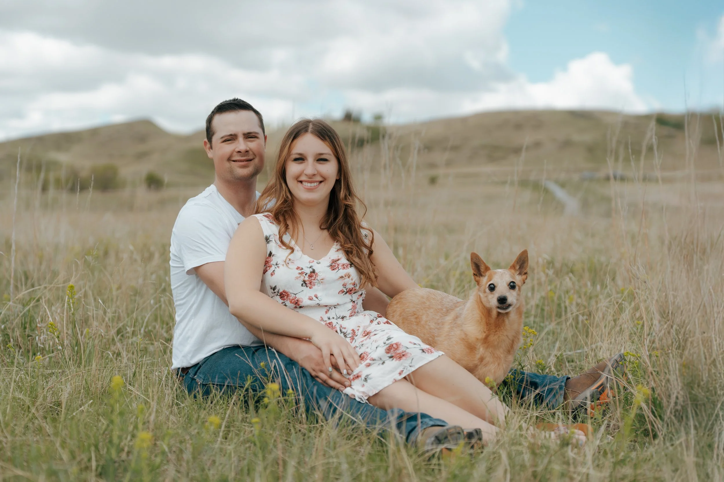 A smiling couple sitting on the grass in a field with a small dog, with rolling hills and a partly cloudy sky in the background.