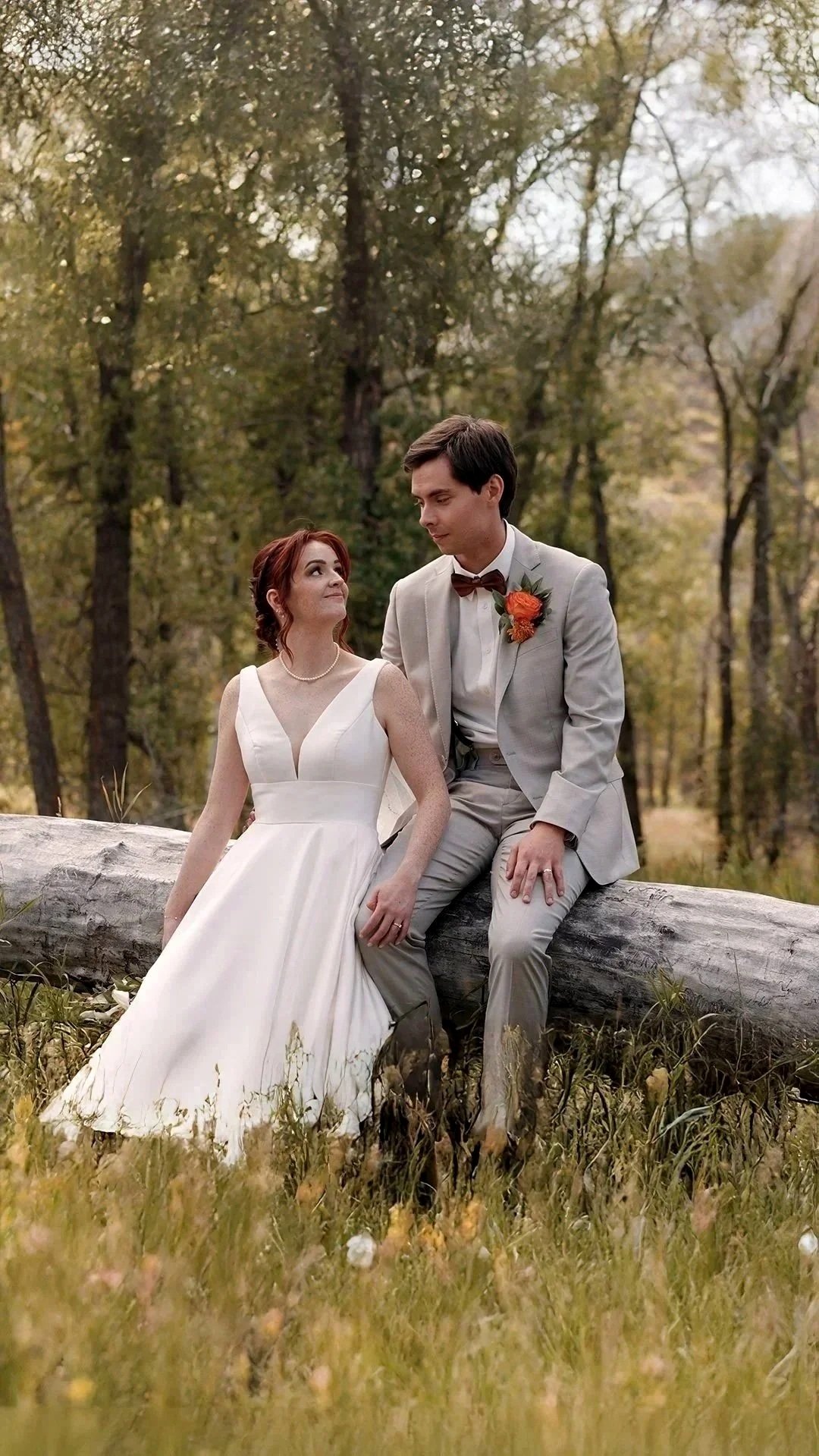 A bride and groom sitting on a fallen log in a forest during autumn, with trees and yellow leaves in the background. The bride is wearing a white wedding dress and the groom is dressed in a light gray suit with a brown bow tie and orange boutonniere.
