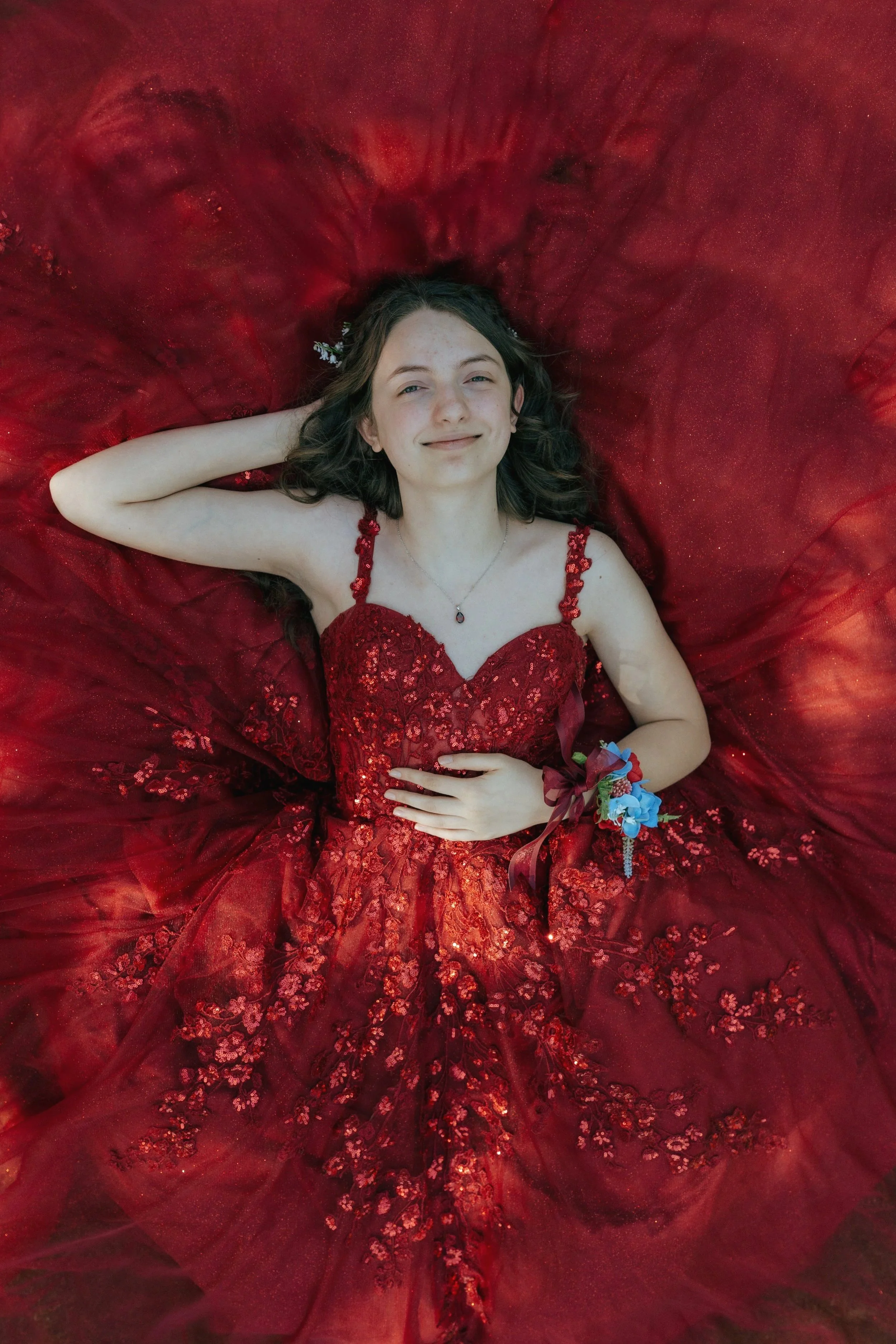 Young girl in an elegant red dress lying on a flowing red fabric, smiling with one arm behind her head and the other resting on her stomach, wearing a corsage on her wrist and a necklace.