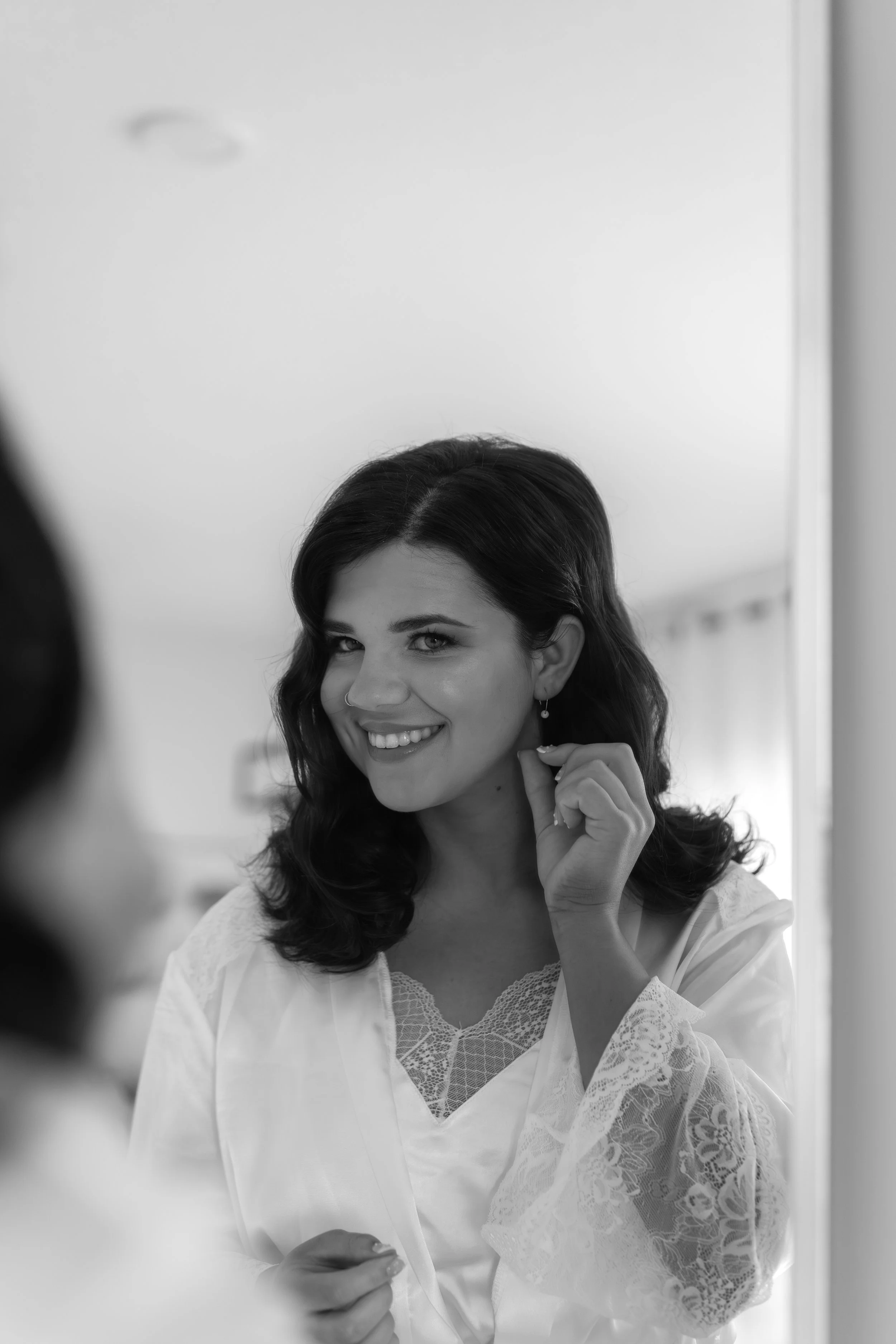 A woman with dark hair smiling at herself in a mirror, adjusting an earring, wearing a silky robe with lace details.
