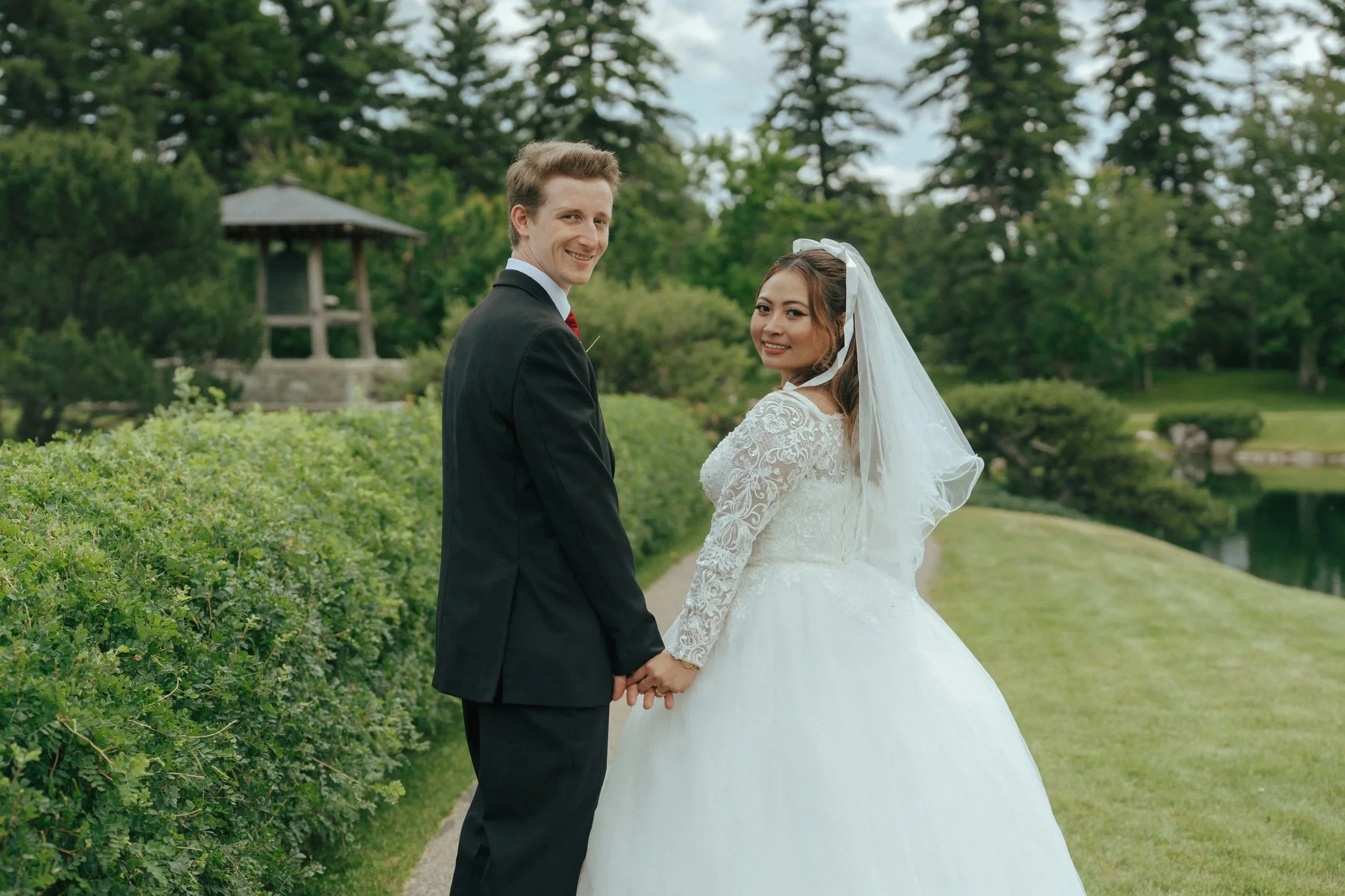 A newlywed couple holding hands and smiling outdoors in a lush green park with trees and a pond in the background.