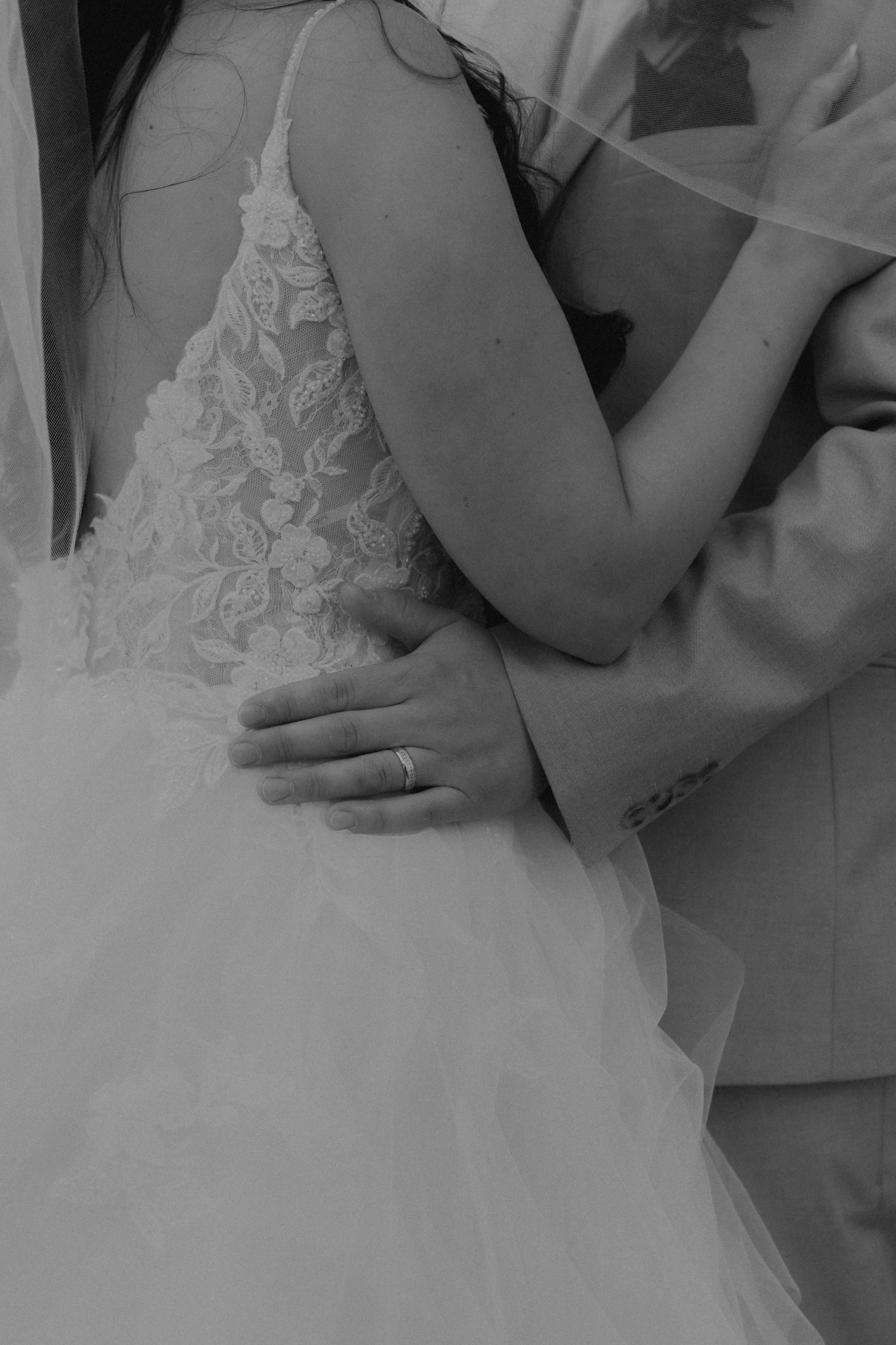 Close-up of a bride and groom embracing, bride wearing a lace wedding dress, groom in a suit, a wedding ring visible on the bride's finger.