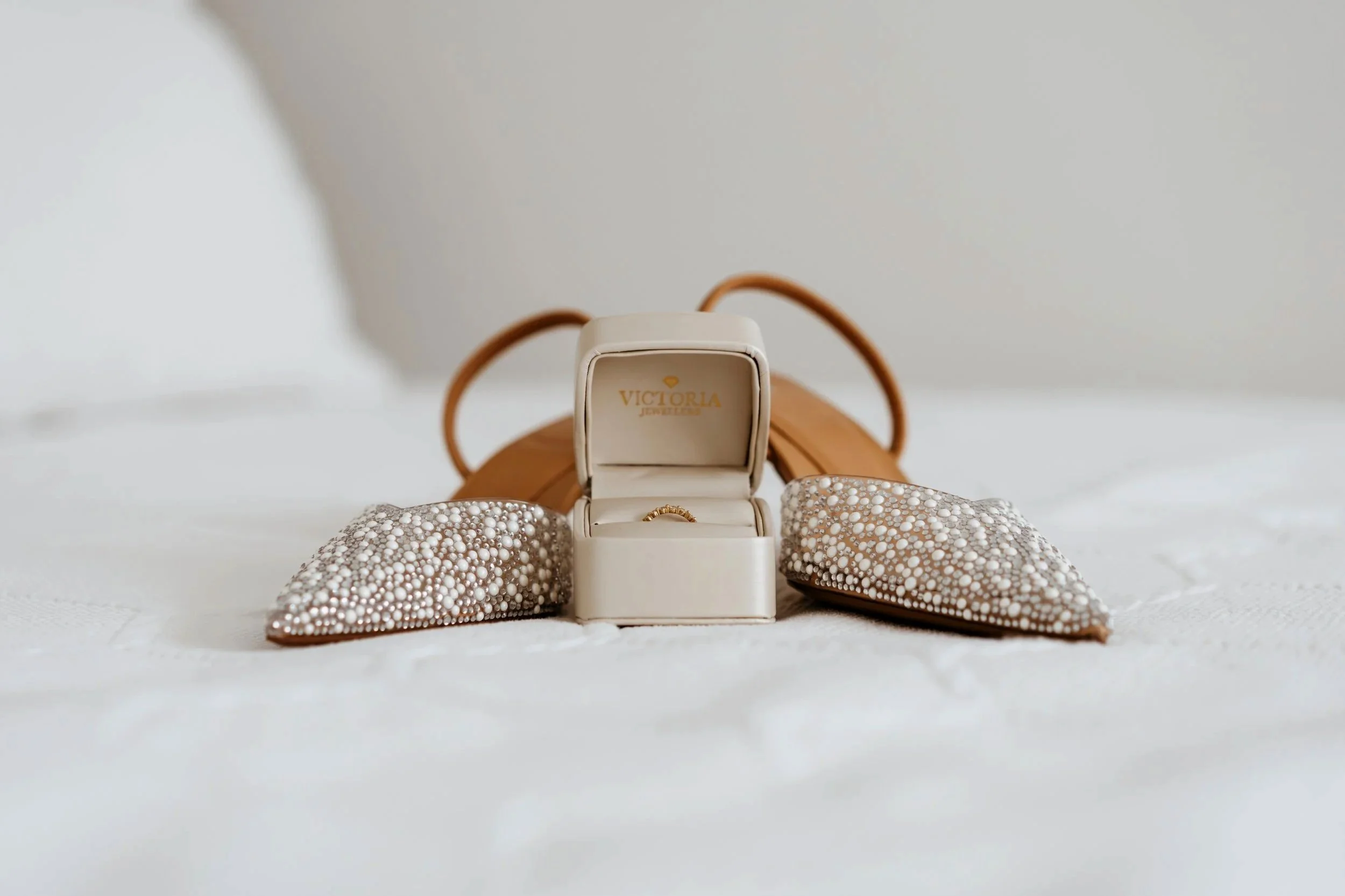 An open jewelry box with a gold ring inside, surrounded by pearl-embellished accessories, with a tan handbag in the background on a white surface.