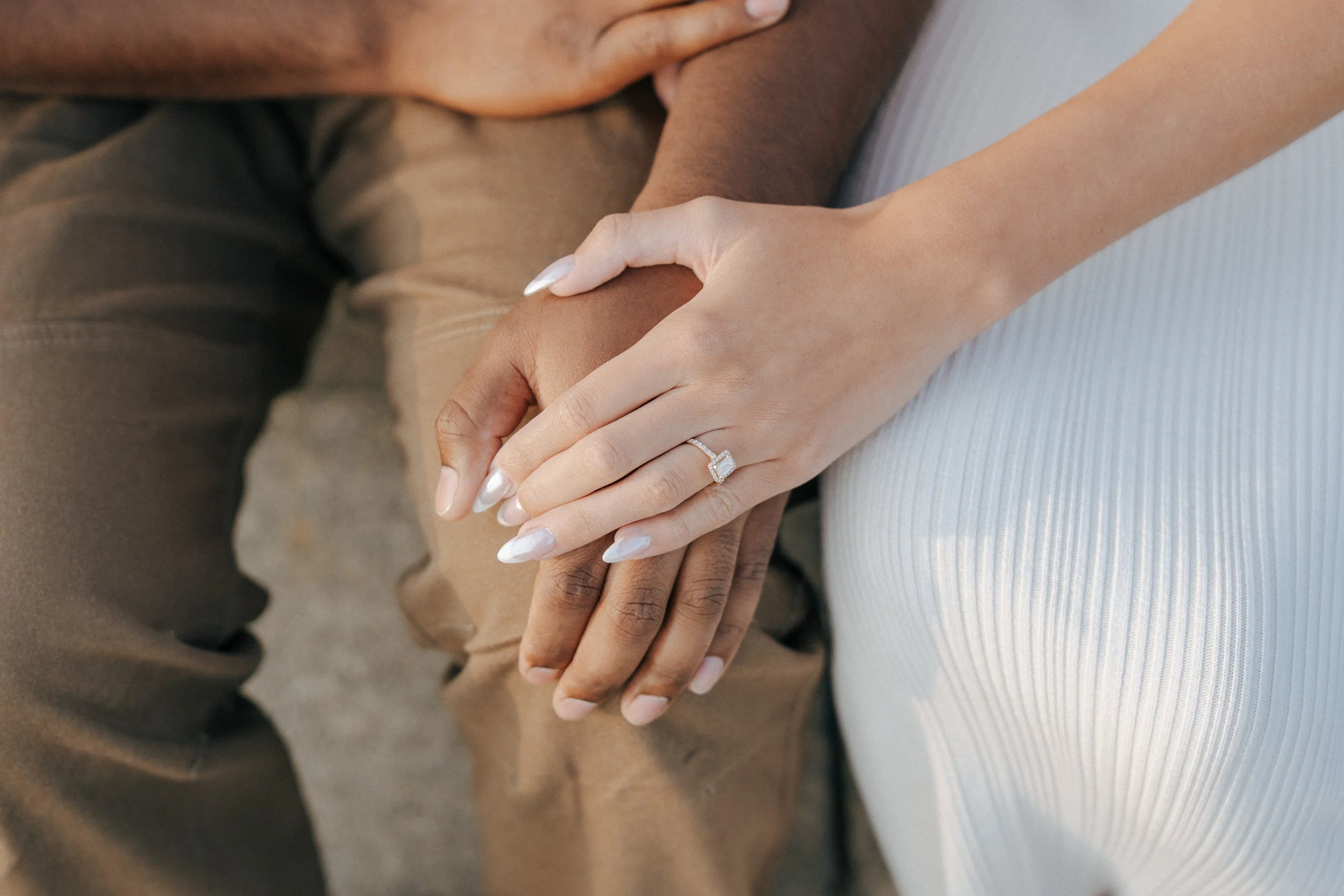 Close-up of a woman’s hand with a diamond engagement ring resting on a man’s hand, with both sitting side by side.