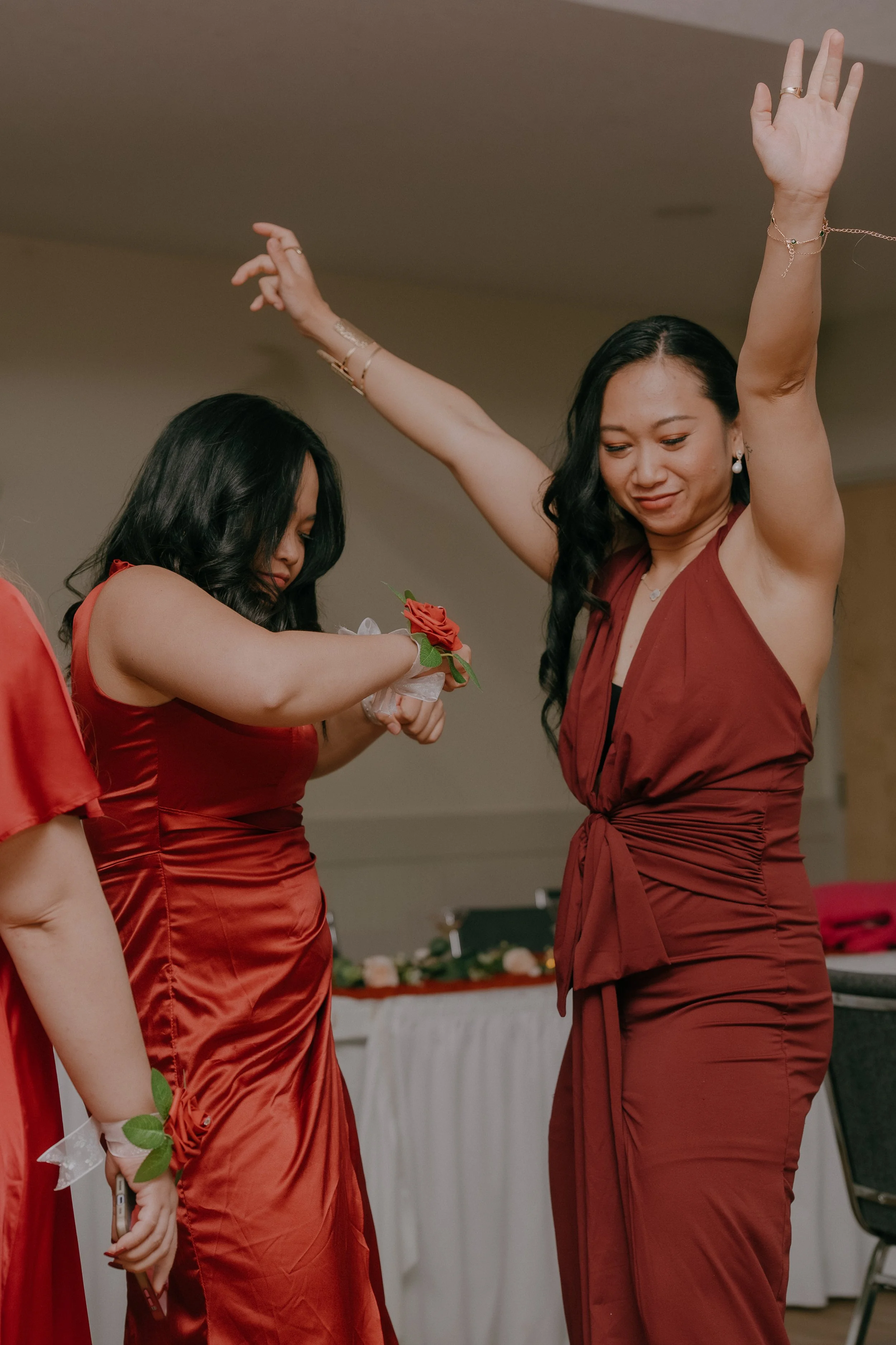 Two women dancing at a formal event, both wearing red dresses. One woman has her arm raised, and the other is looking down, holding a wrist corsage with a red flower.