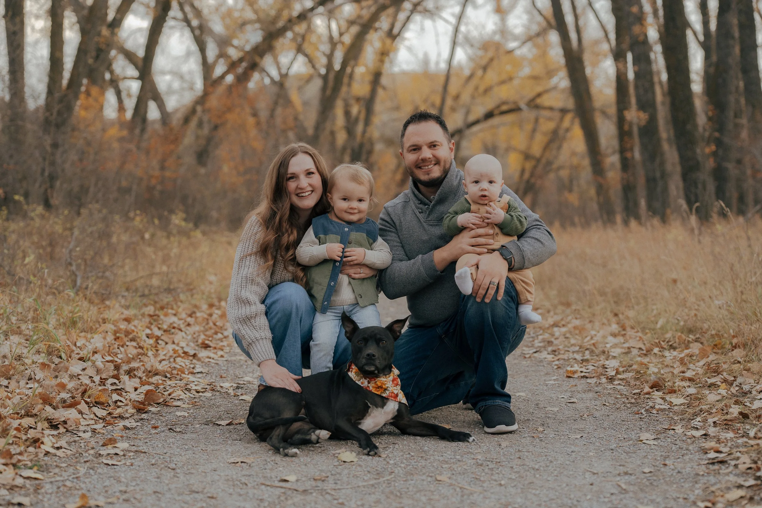 A family of four and a dog posing outdoors on a trail in autumn, surrounded by trees with fall foliage. The mother, father, and two young children are smiling, and the dog is lying in front of them.